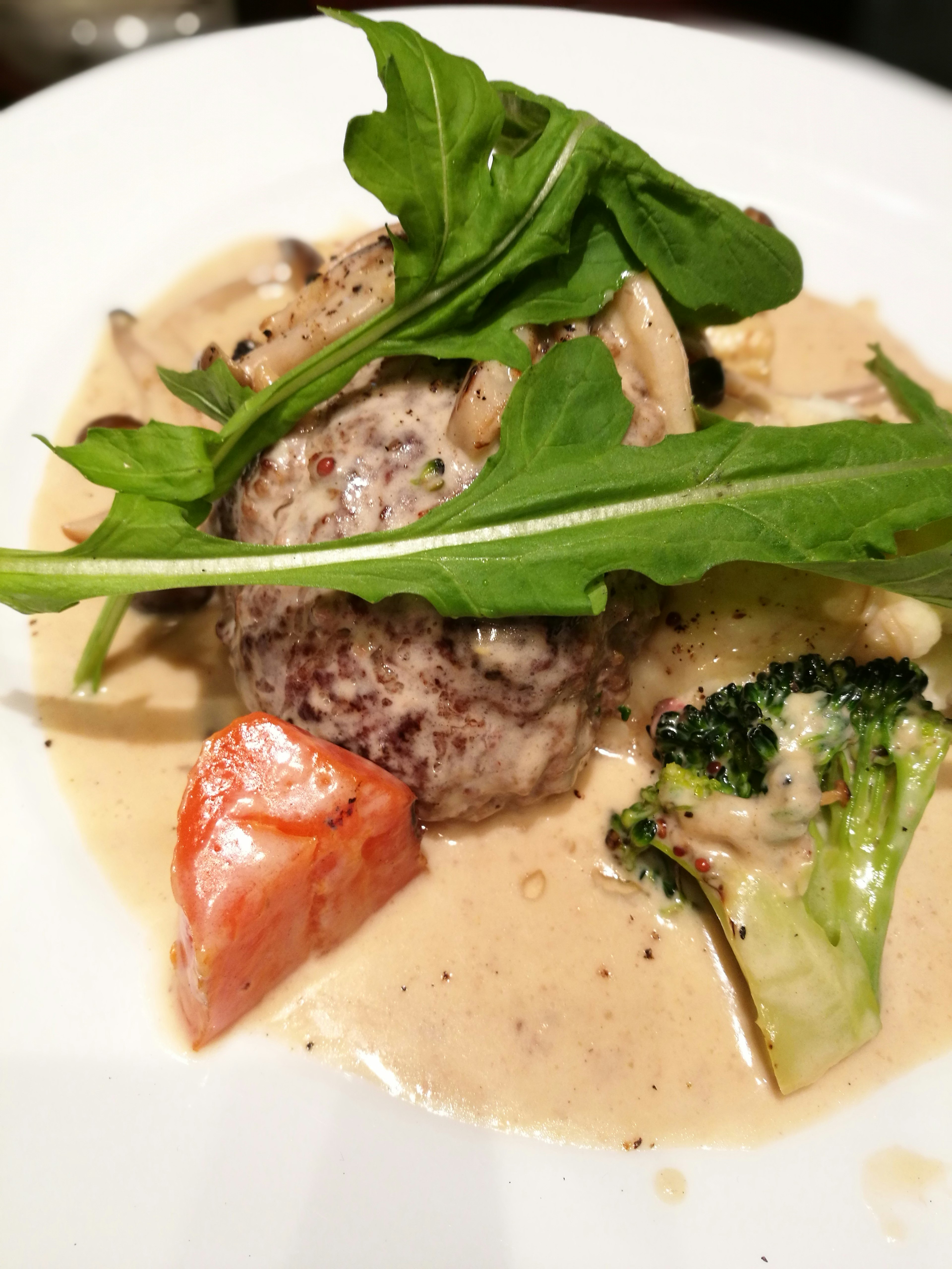 Dish featuring meat patty topped with greens and served in creamy sauce with vegetables