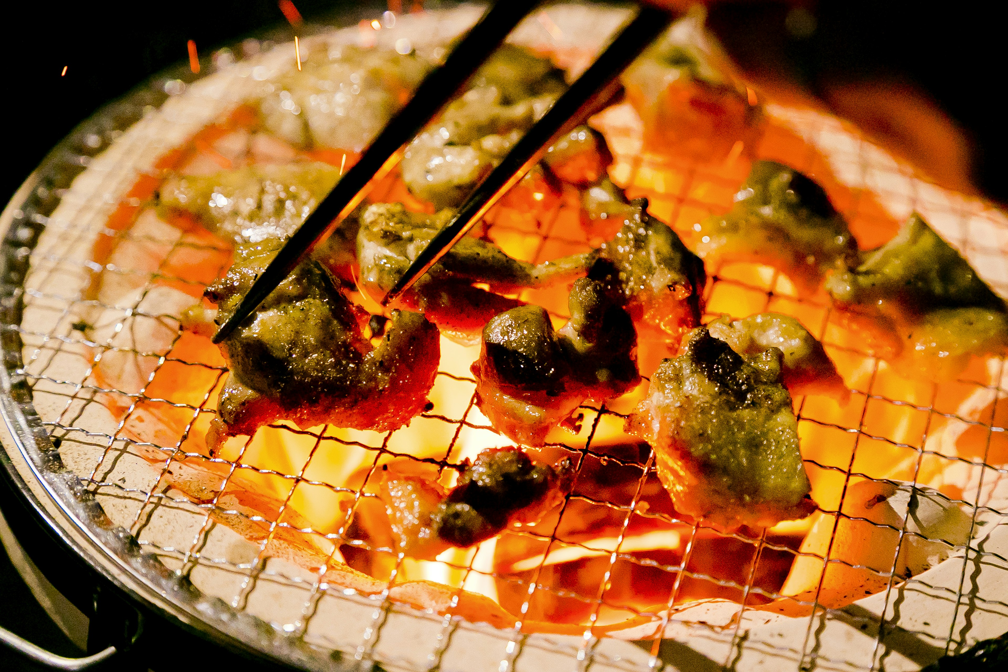 Chunks of meat grilling over charcoal with chopsticks picking them up