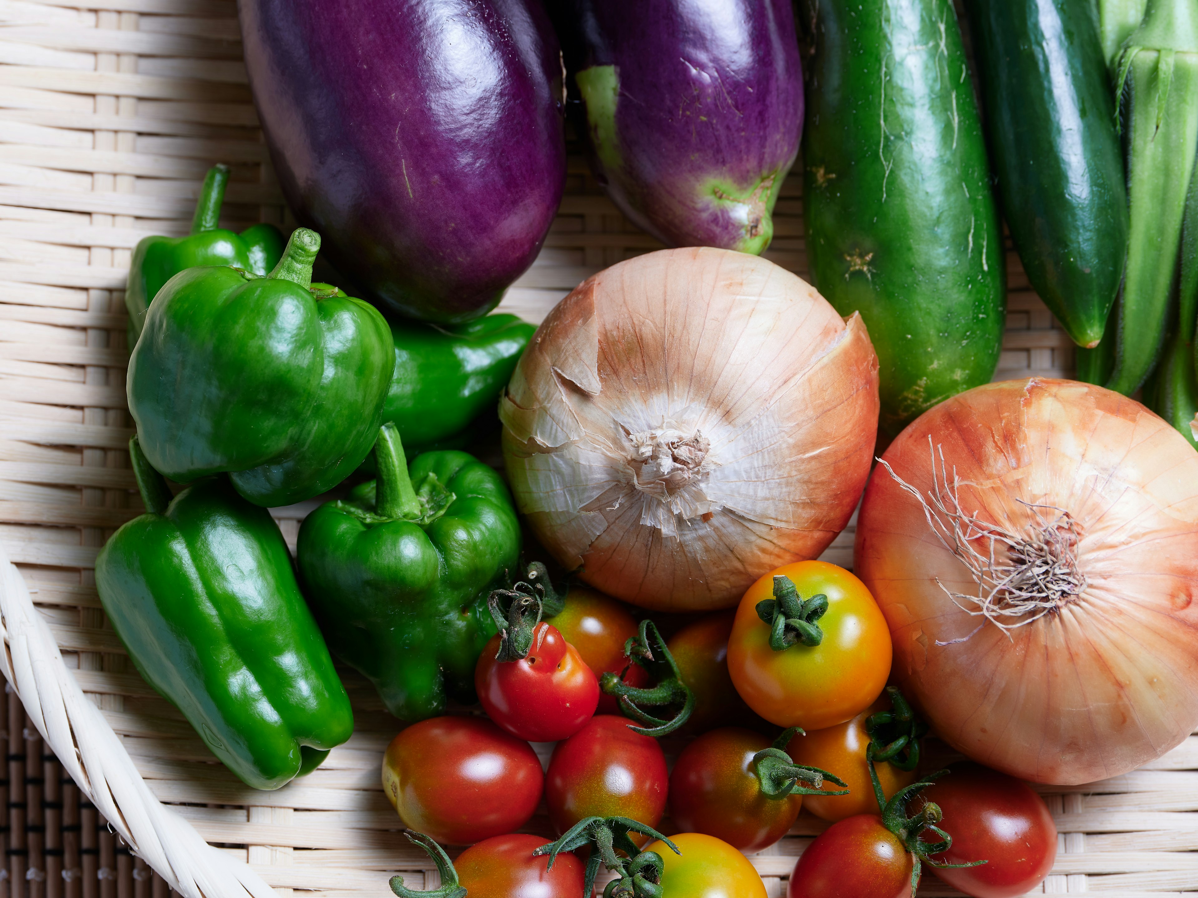 A basket filled with colorful vegetables including eggplants green peppers onions and cherry tomatoes