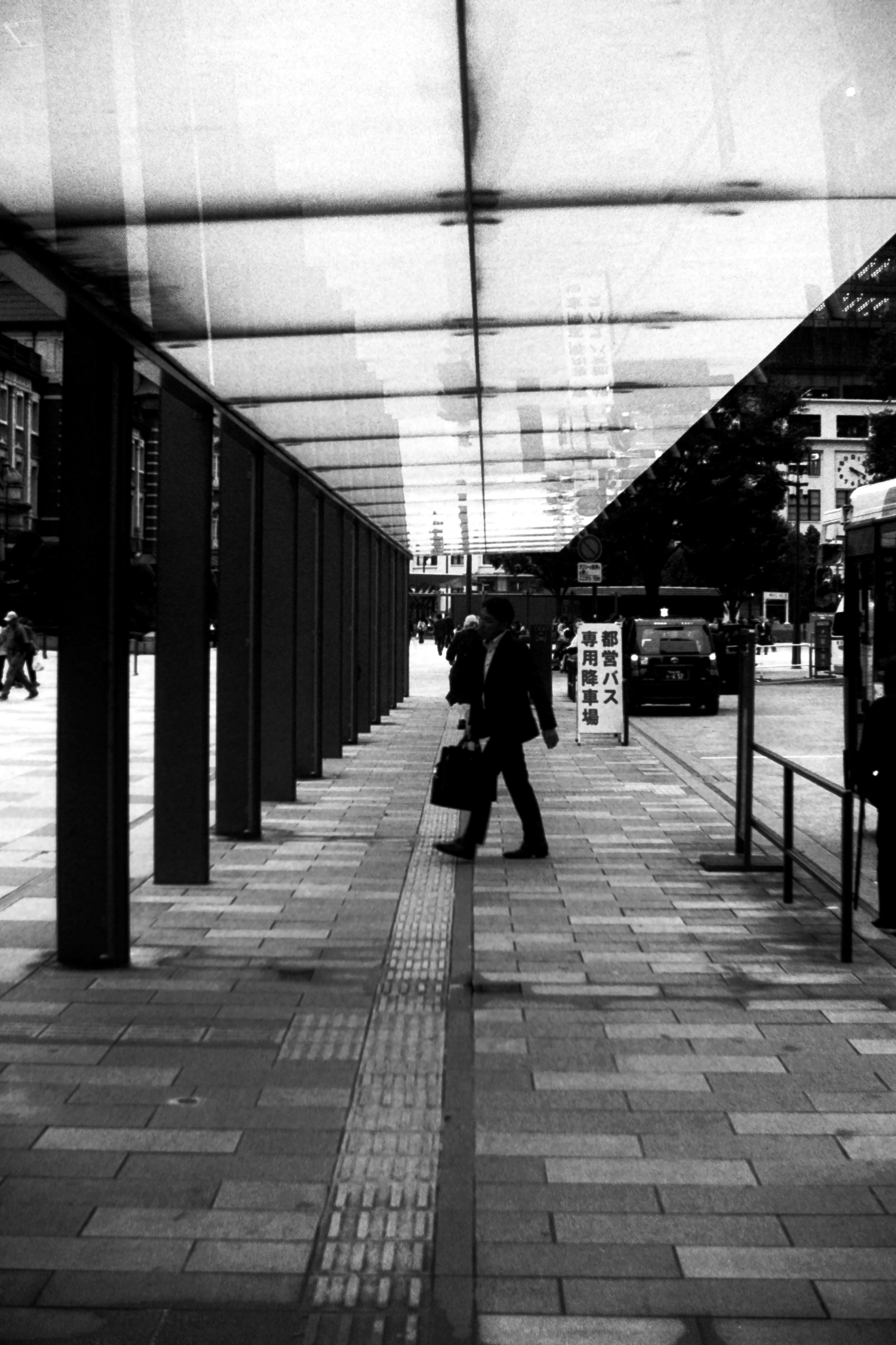 Une personne marchant sur un trottoir sous un toit transparent dans un cadre urbain en noir et blanc