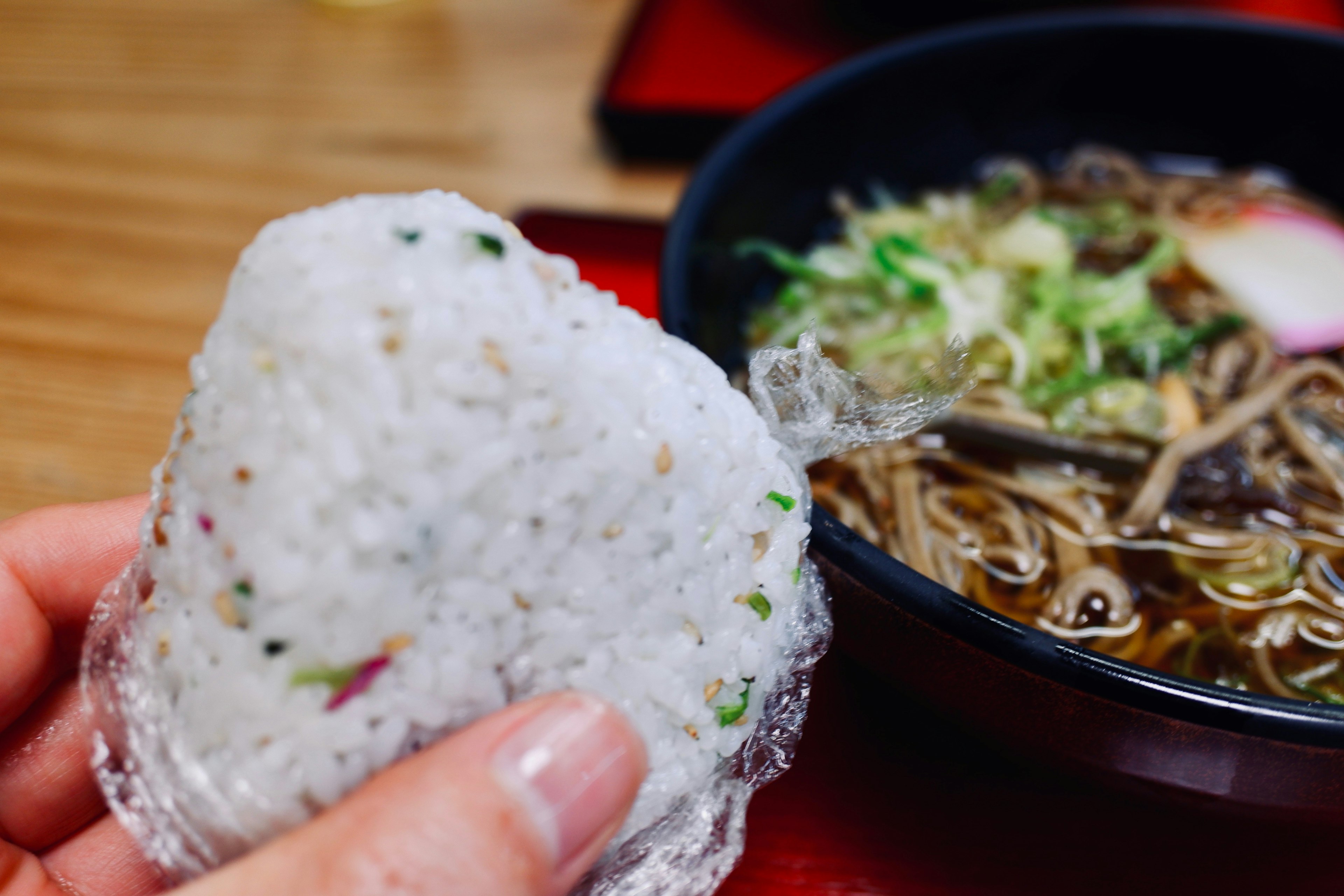 A hand holding a rice ball with a bowl of ramen in the background