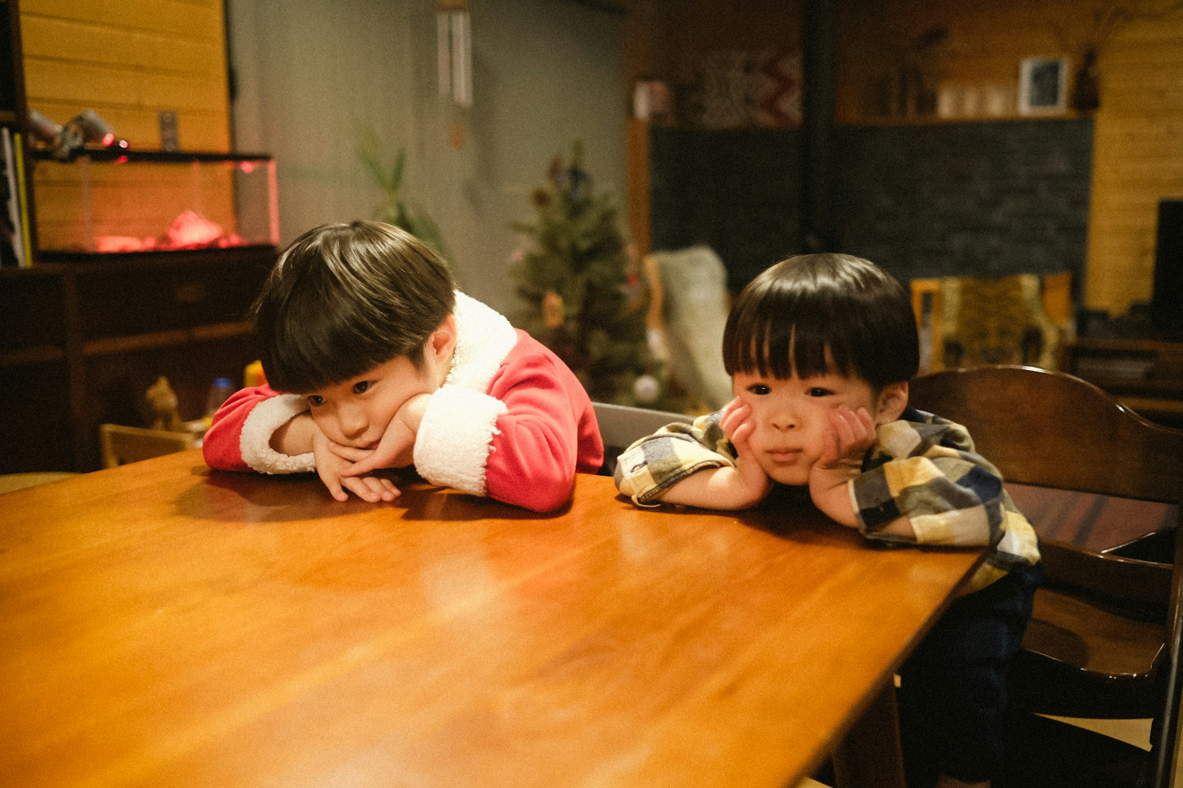 Child in Santa costume and child in checkered shirt resting their heads on a table