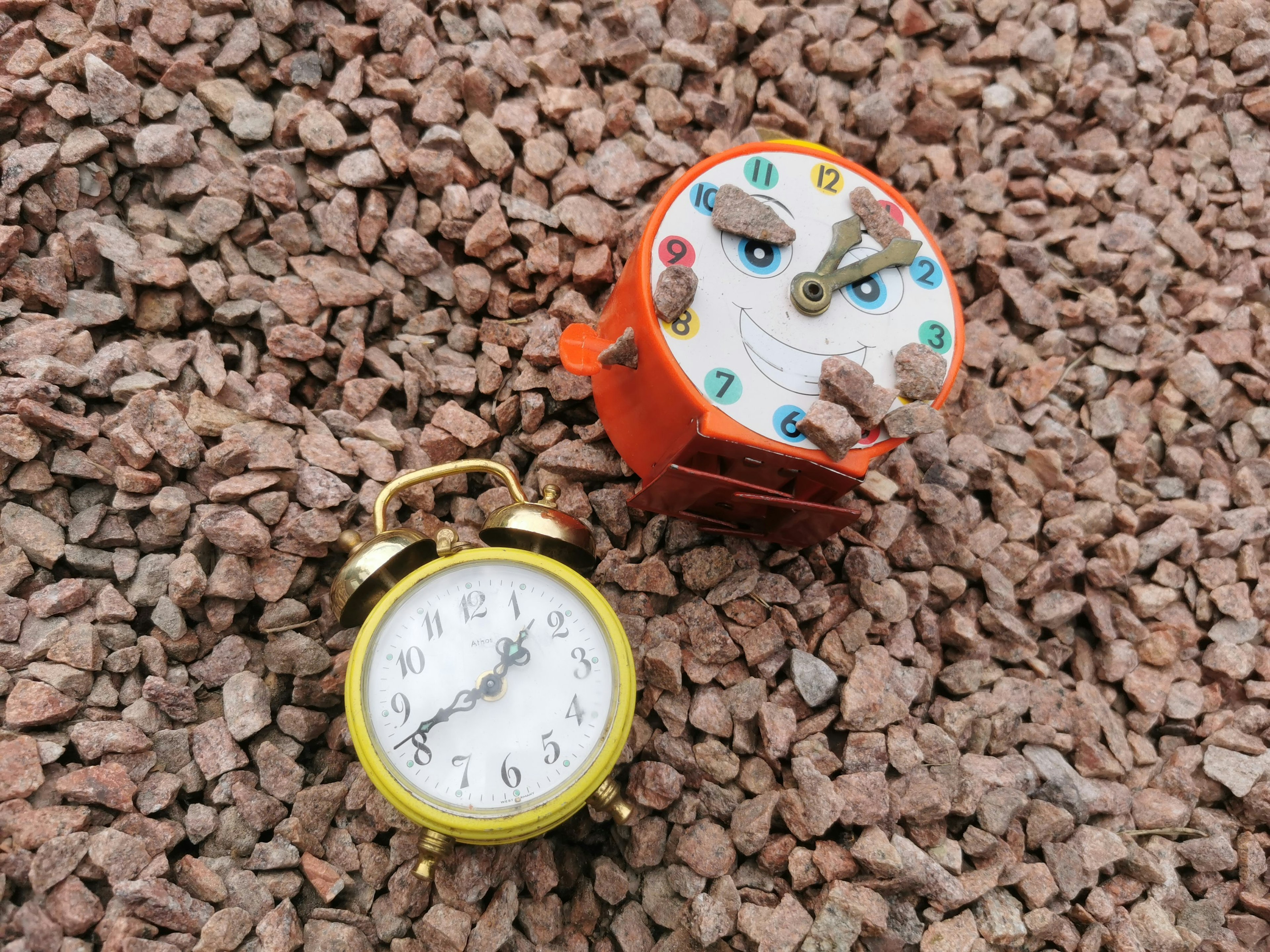 An orange children's clock and a yellow alarm clock placed on gravel