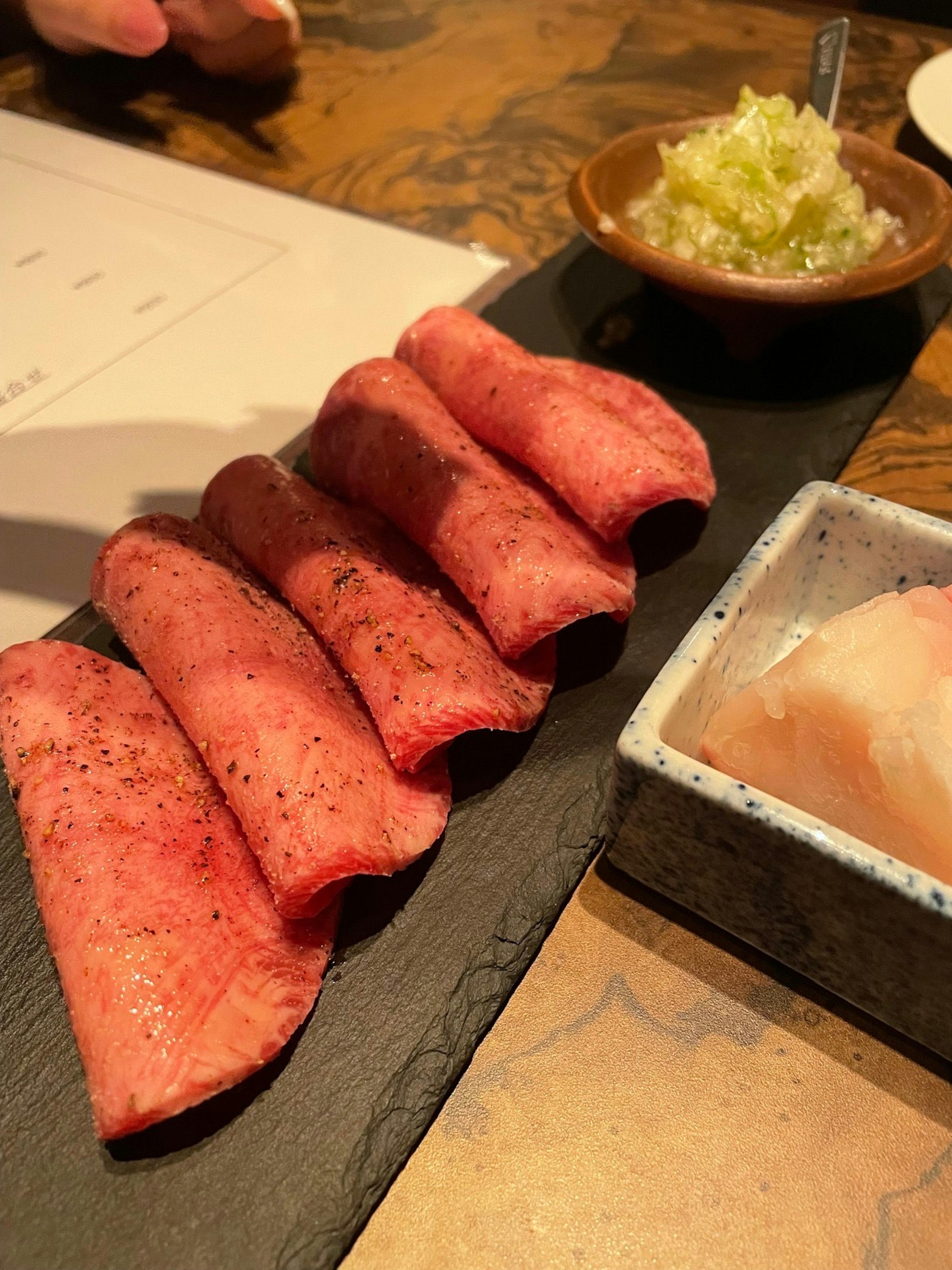 Five slices of marbled meat on a black plate accompanied by a small bowl of garnishes