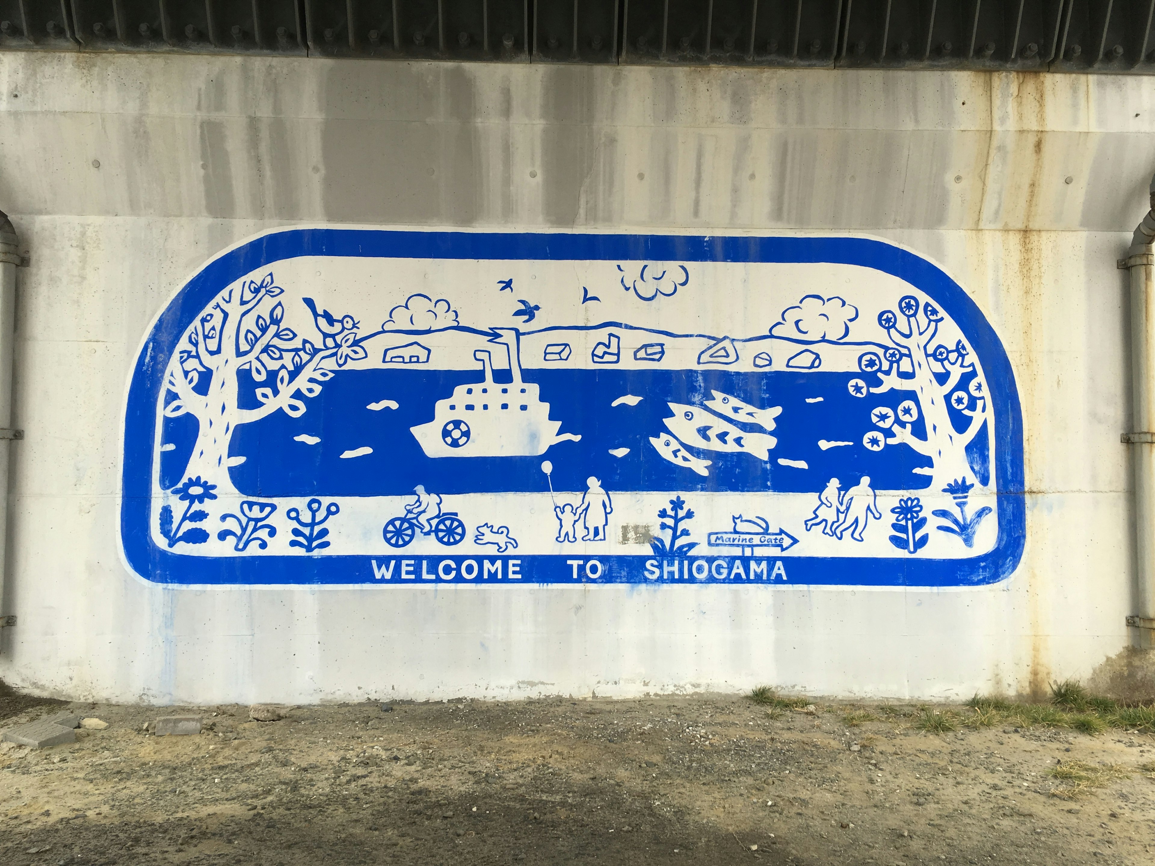 Mural biru dan putih yang menggambarkan pemandangan dengan perahu, sepeda, dan pohon dengan teks "WELCOME TO SHIOGAMA" yang mencolok