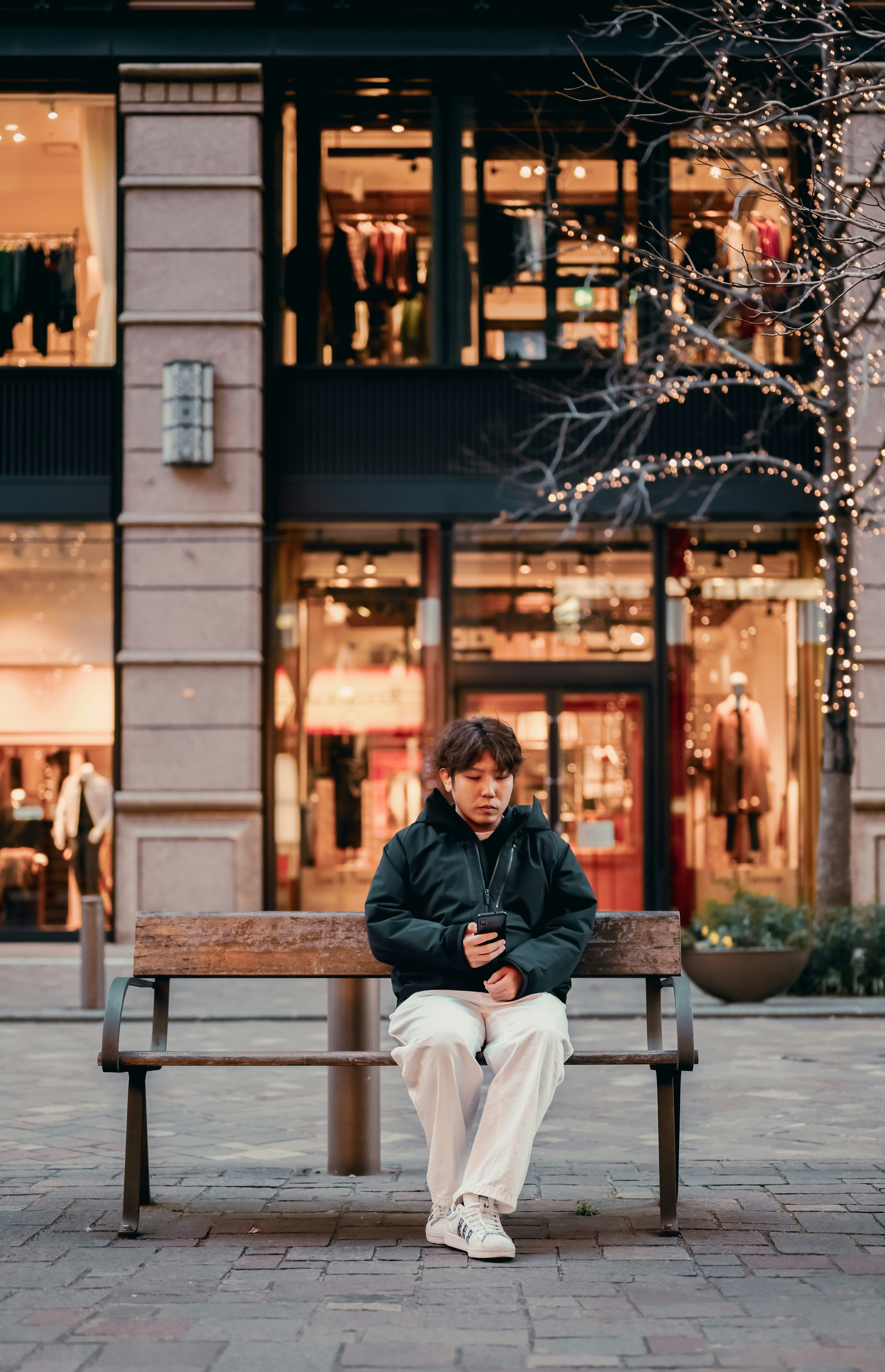 Jeune homme assis sur un banc portant un manteau noir et un pantalon blanc devant un magasin