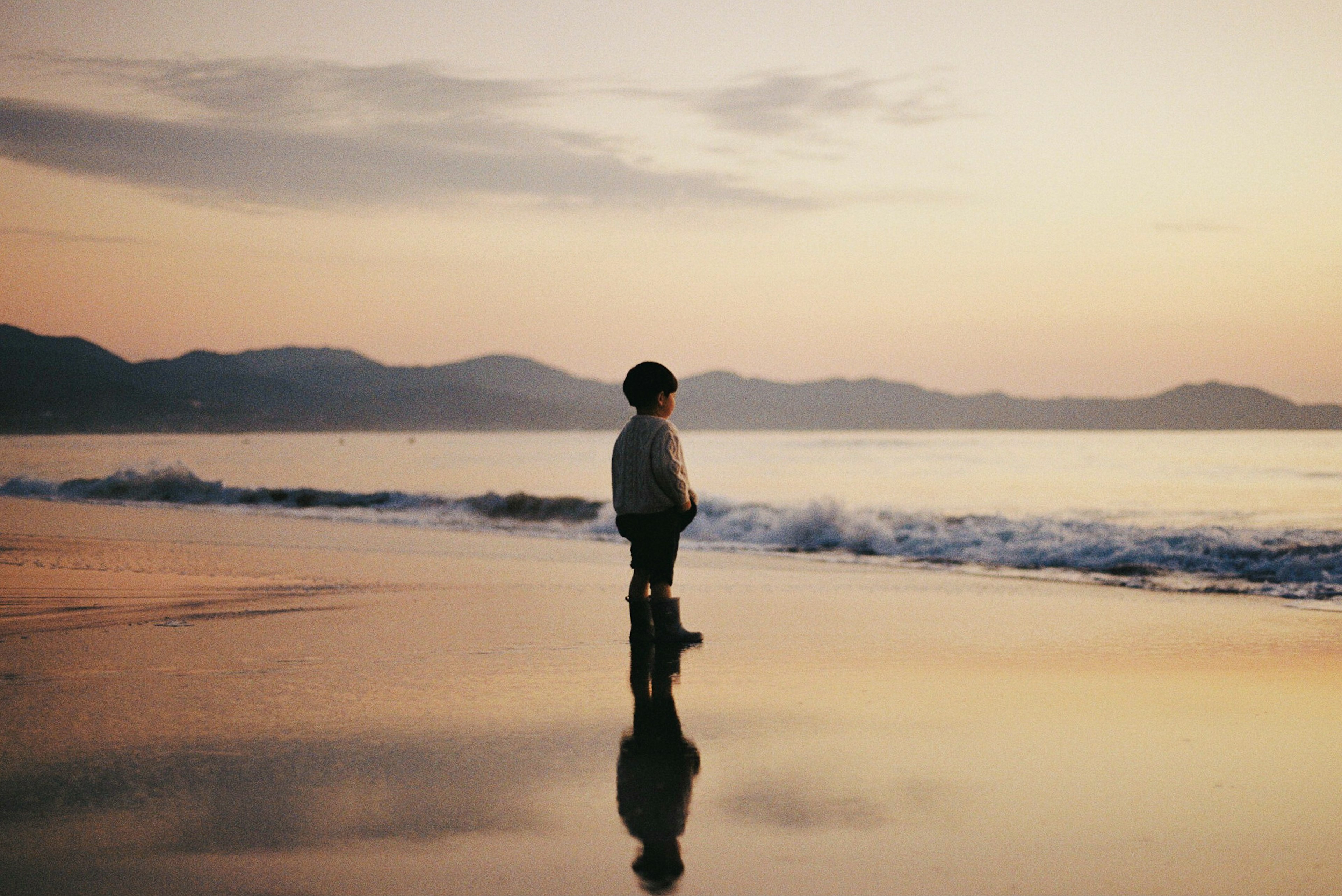 Silhouette eines Jungen, der am Strand mit Sonnenuntergang und Wellenreflexion steht