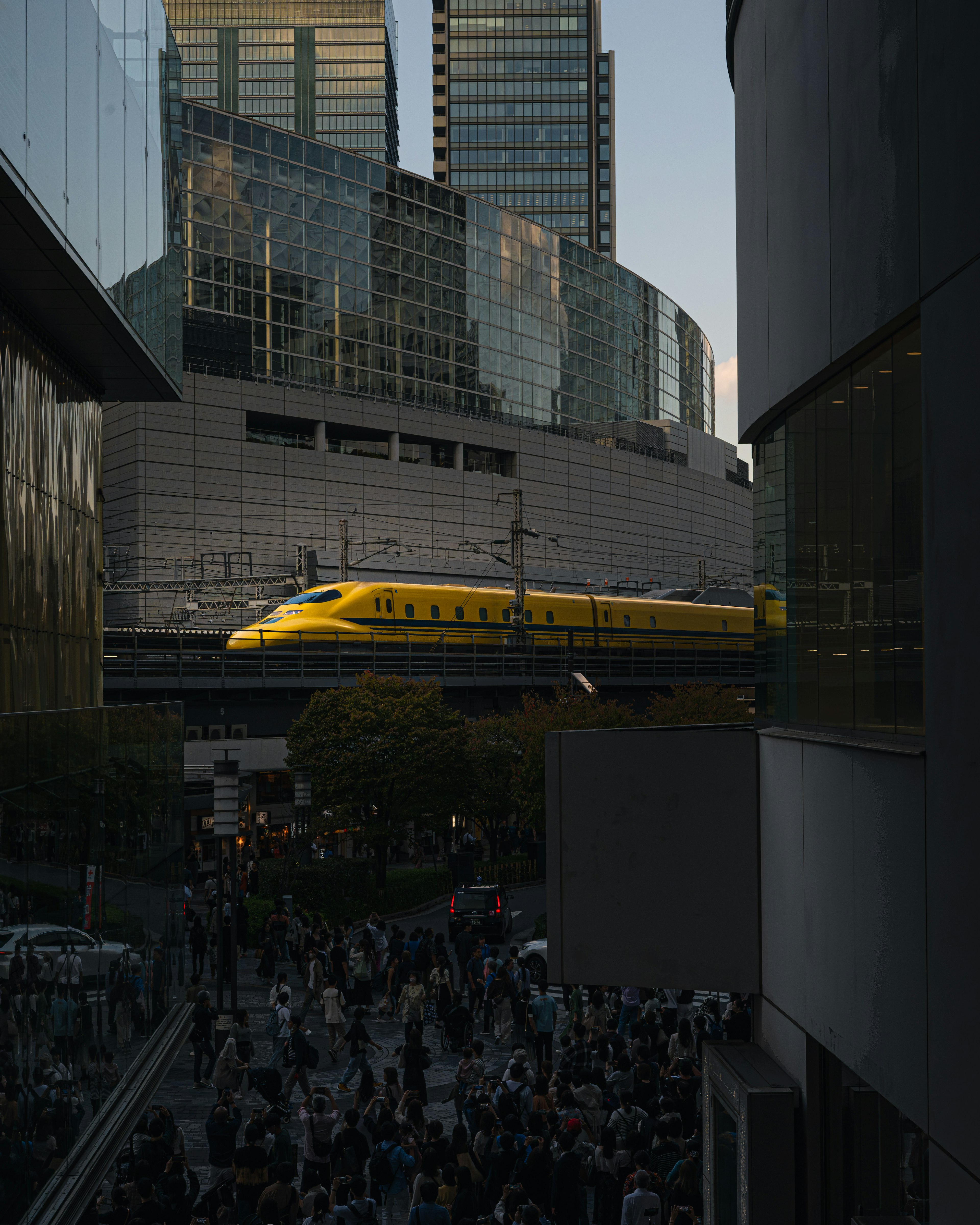 Yellow Shinkansen train passing through a bustling urban scene at dusk