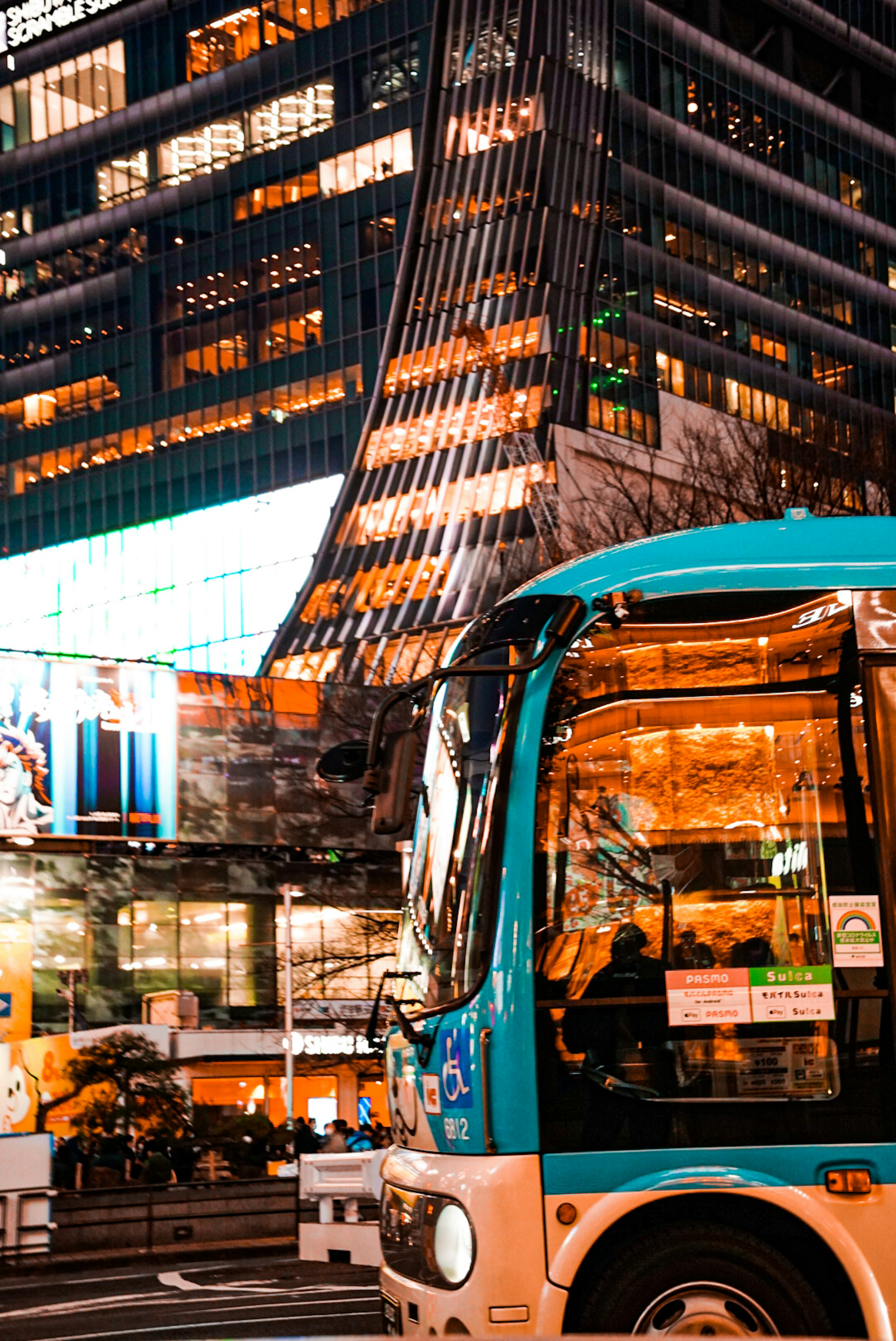 Sebuah bus biru diparkir di pemandangan malam perkotaan yang hidup dengan gedung pencakar langit yang menyala