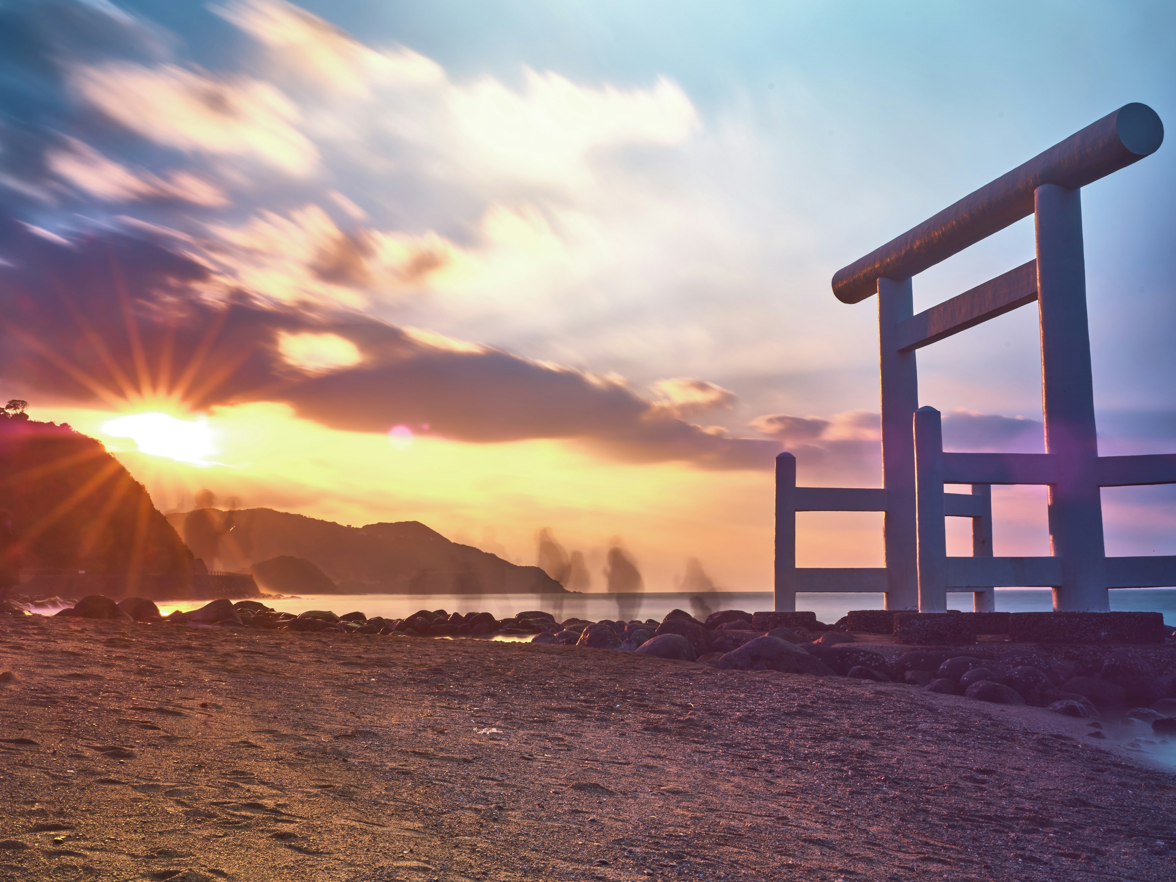 Puerta torii blanca en una playa al atardecer con montañas de fondo