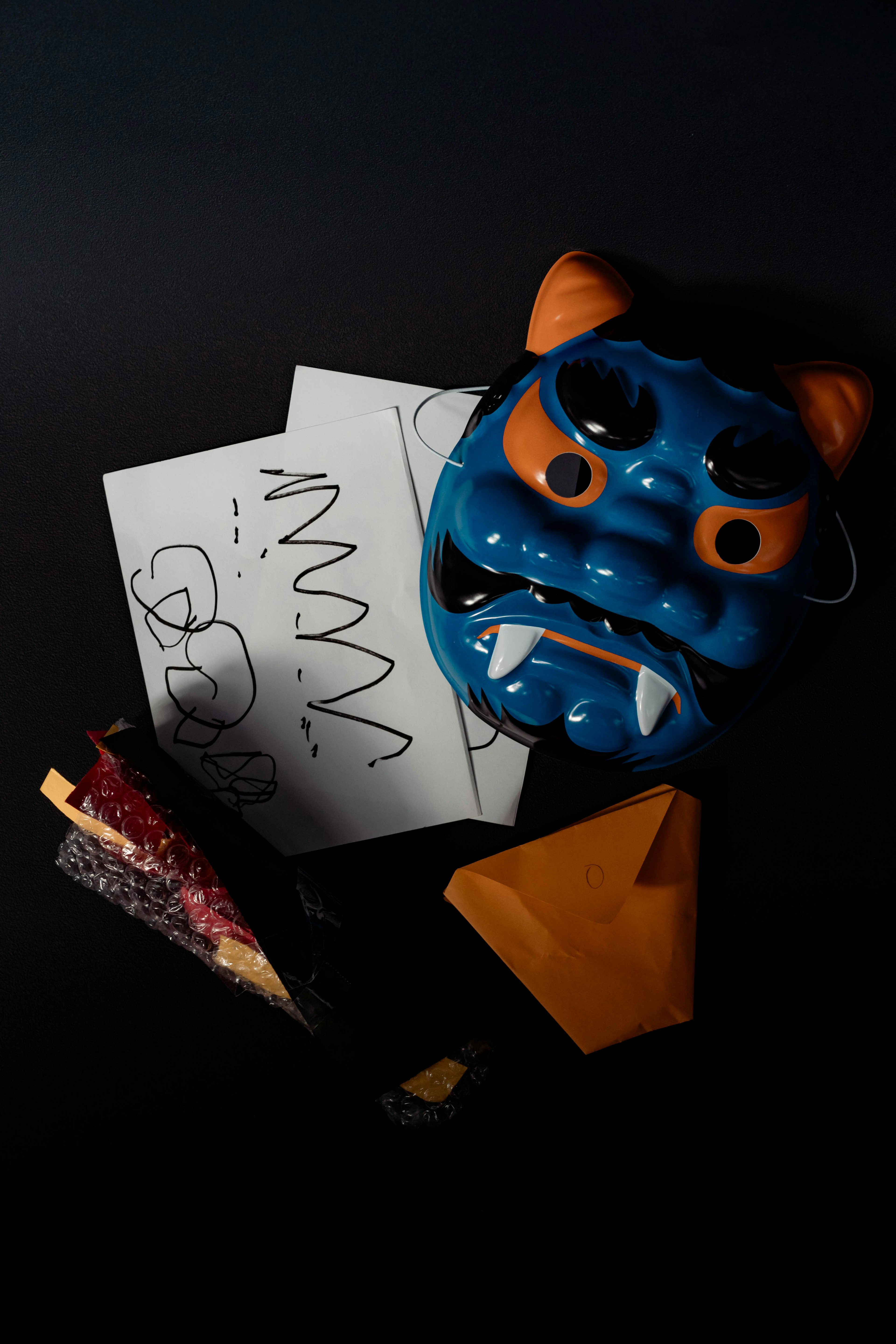 Blue demon mask with orange accents and origami pieces on a black background