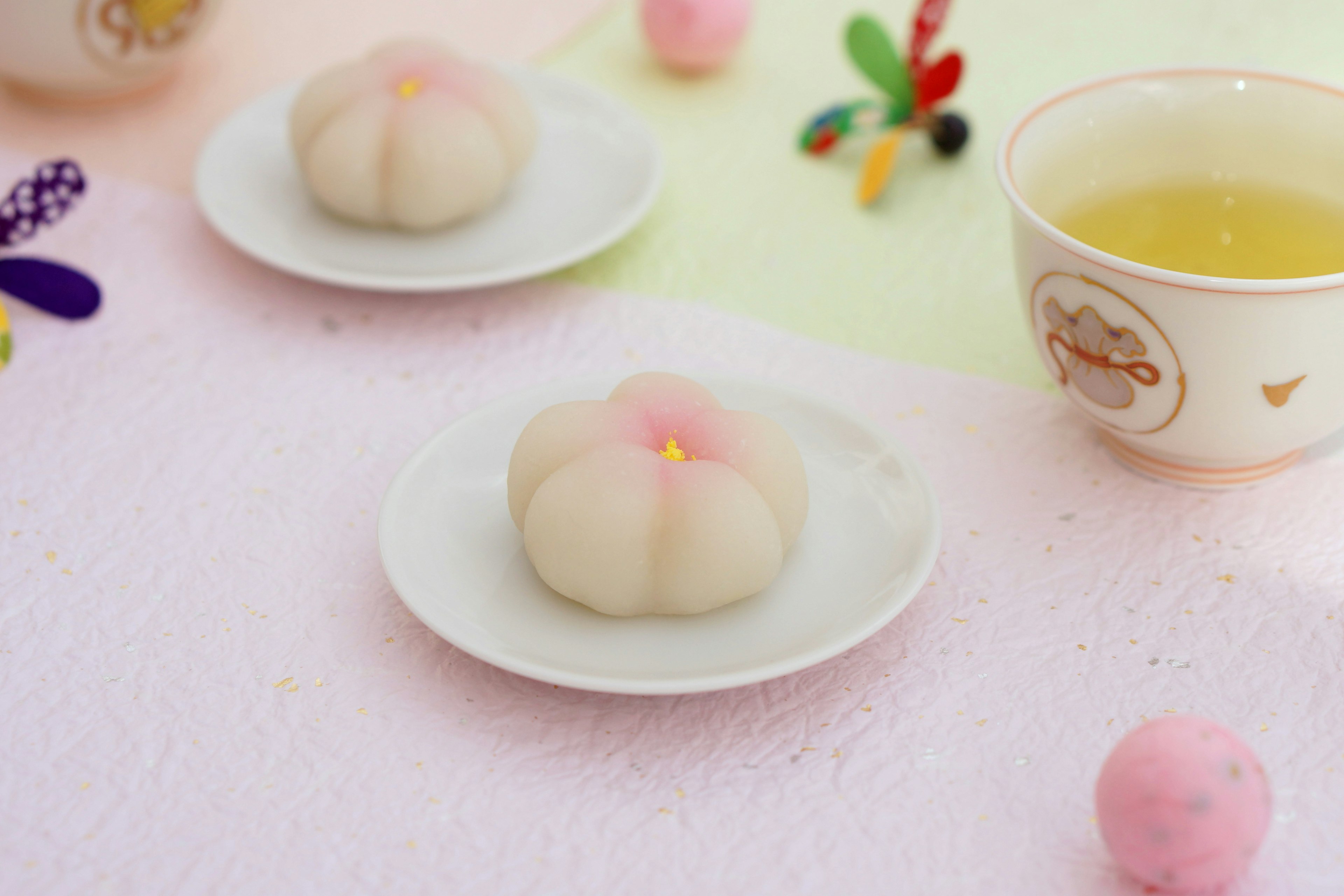 Two flower-shaped wagashi are plated on white dishes with colorful decorations and a cup of tea in the background