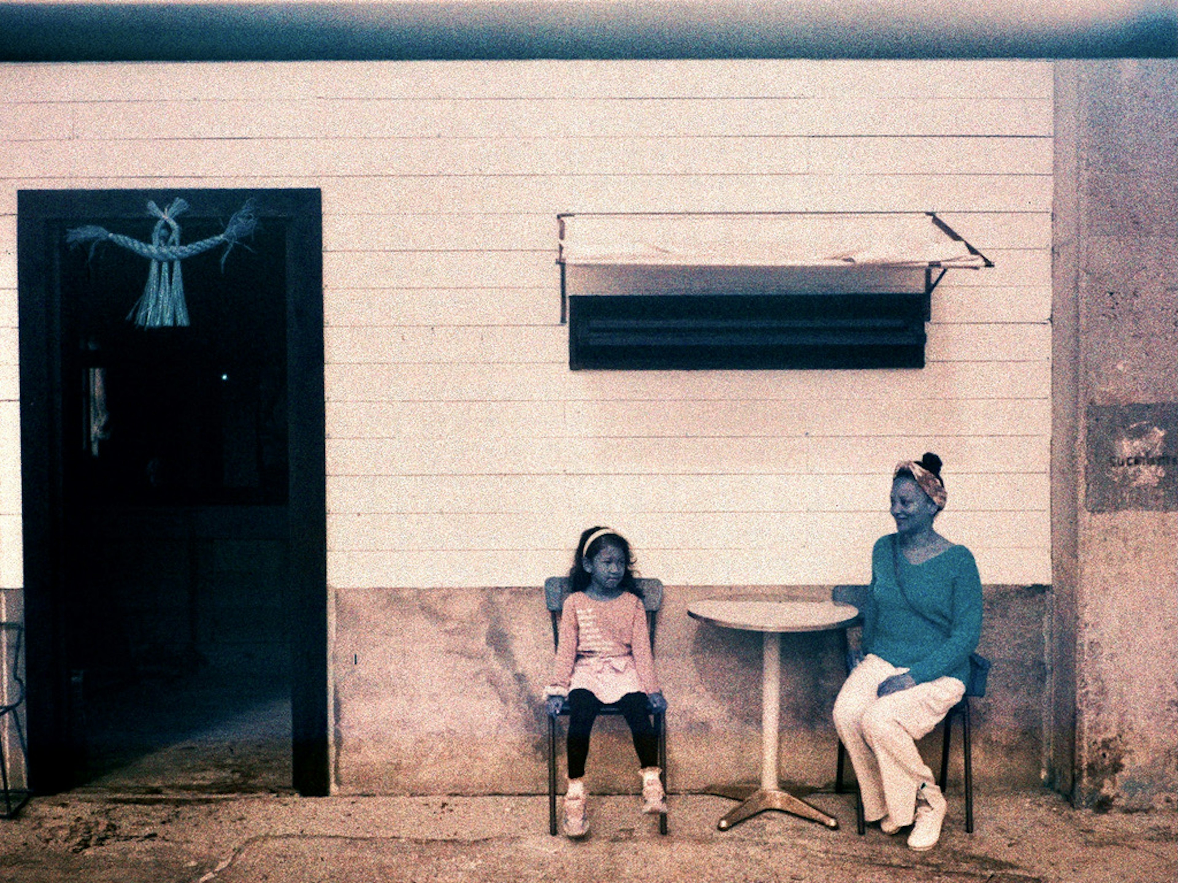 Photo d'une fille et d'une femme assises à l'extérieur devant un mur blanc avec une petite table et des chaises