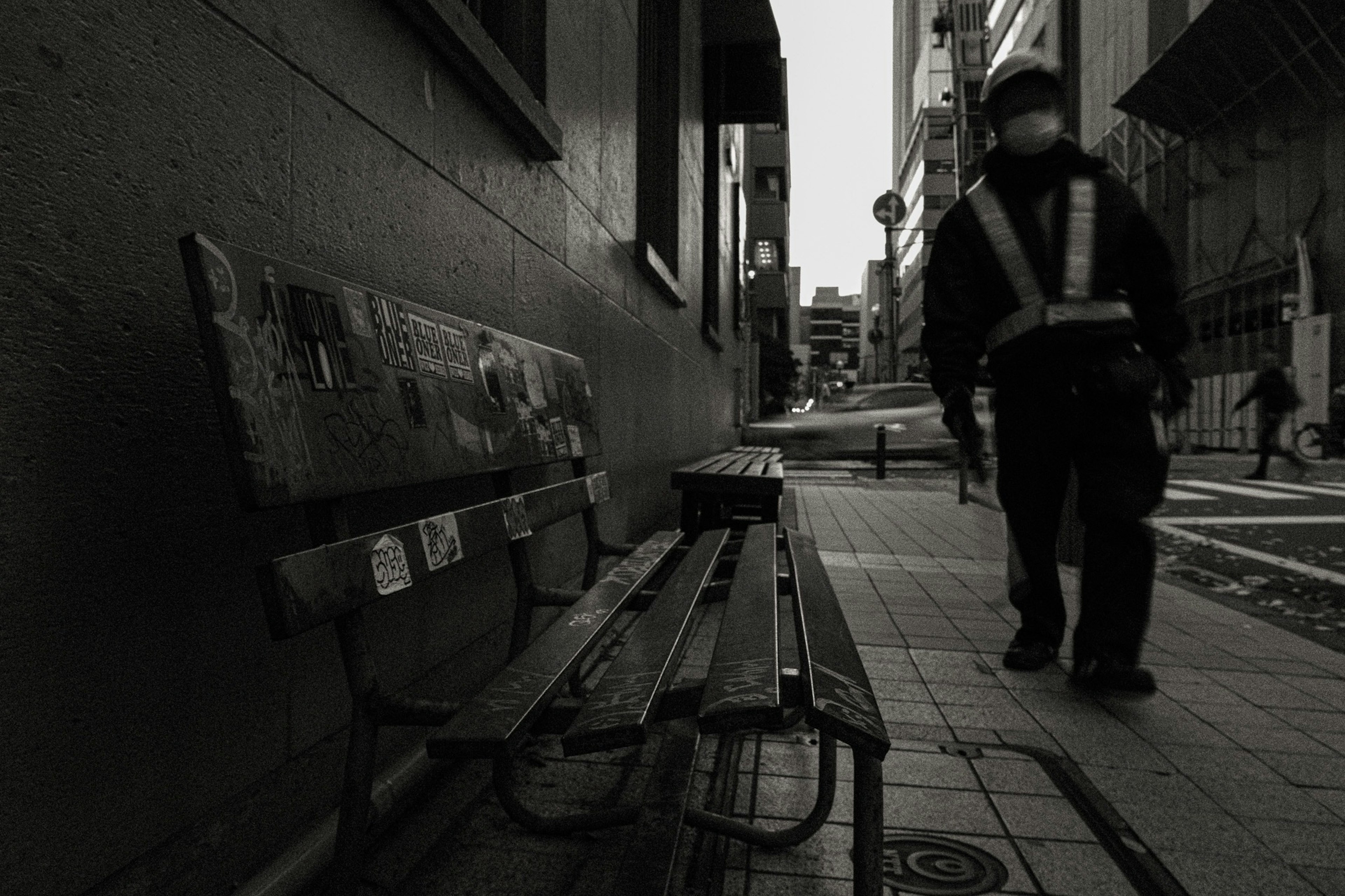 A security guard walking in a black and white urban scene next to a bench