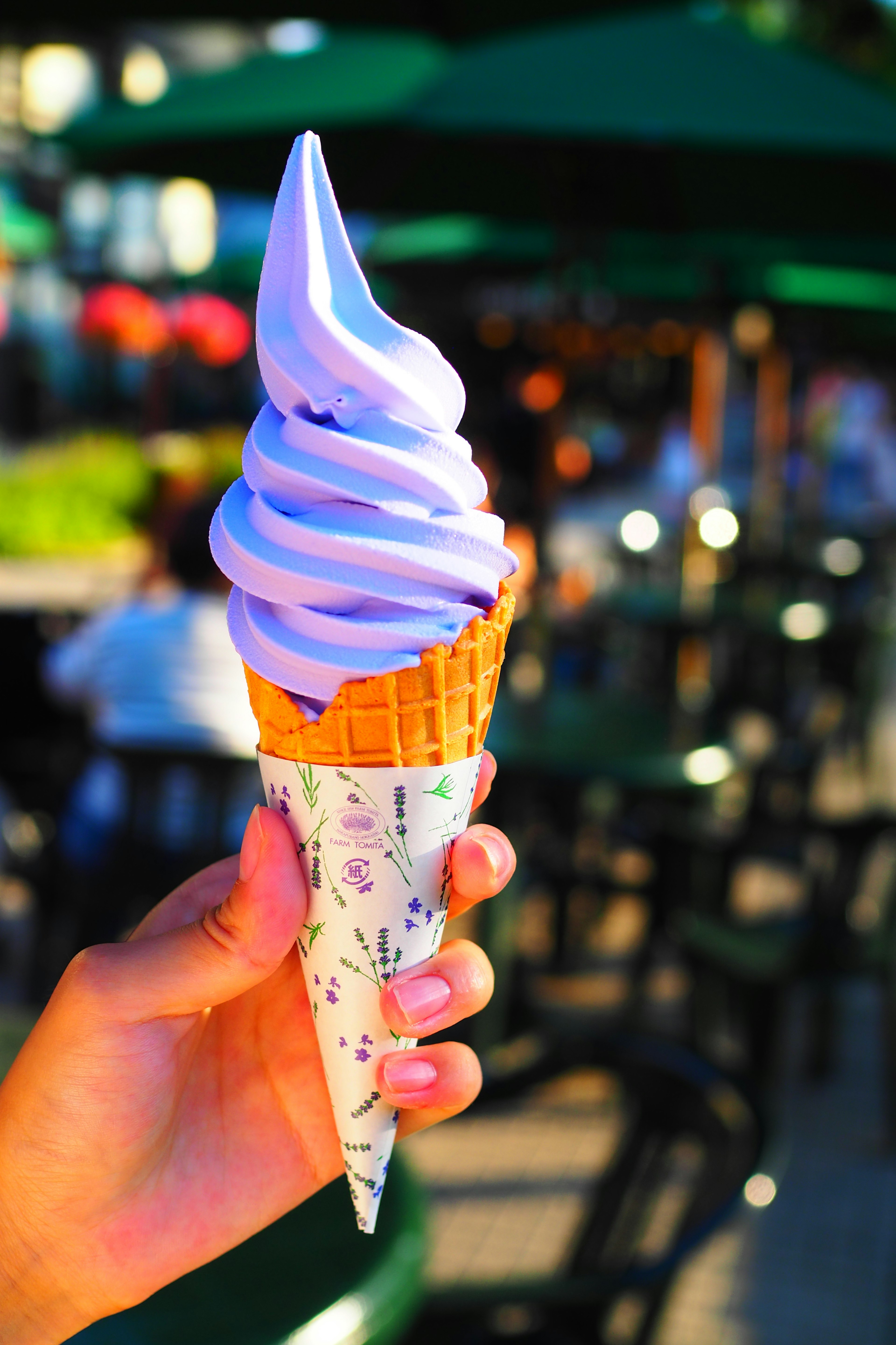 Close-up of a hand holding a purple soft serve ice cream cone with a busy background