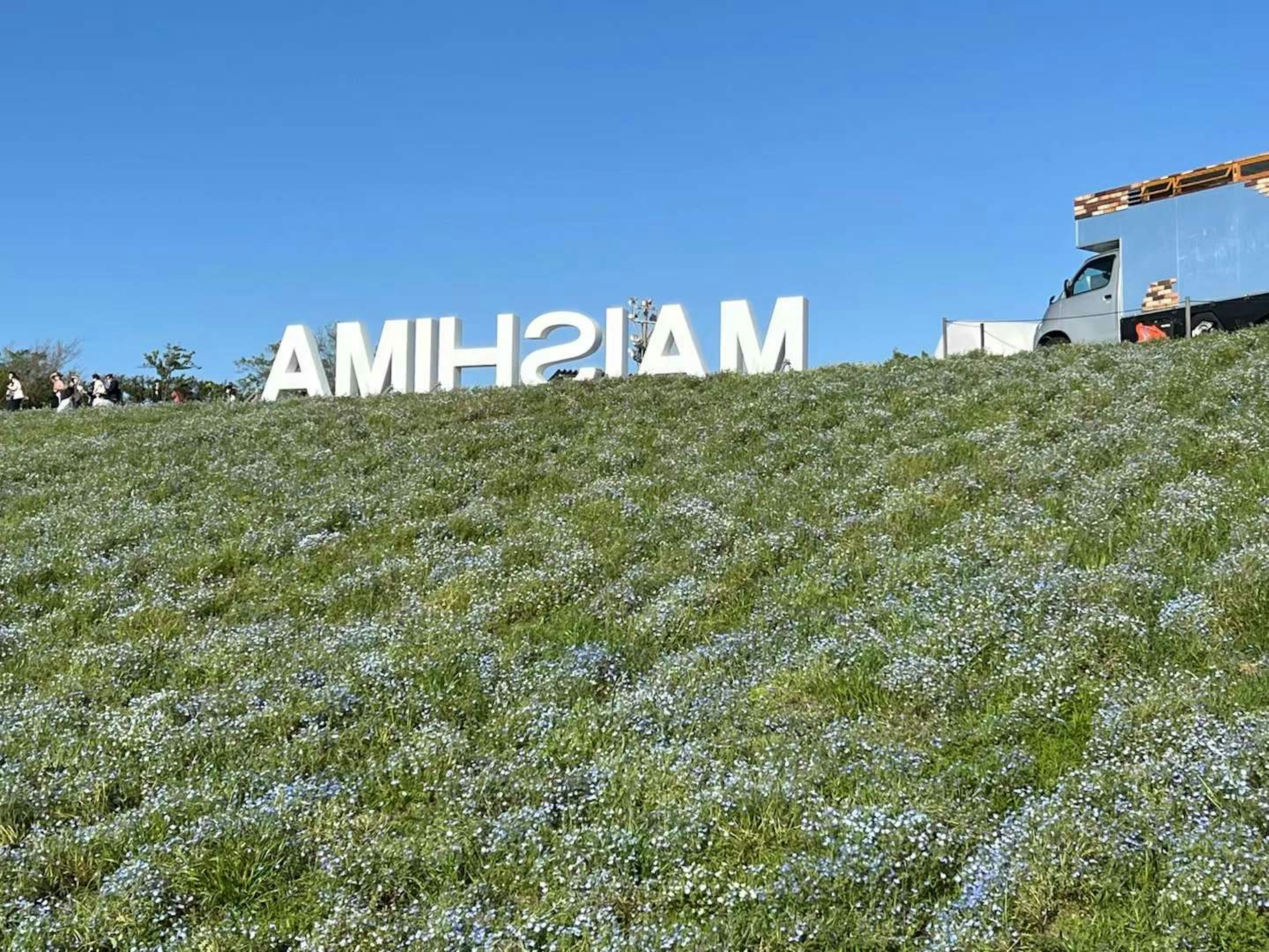 Großes weißes Schild mit der Aufschrift 'MAISHIMA' auf grünem Gras unter blauem Himmel