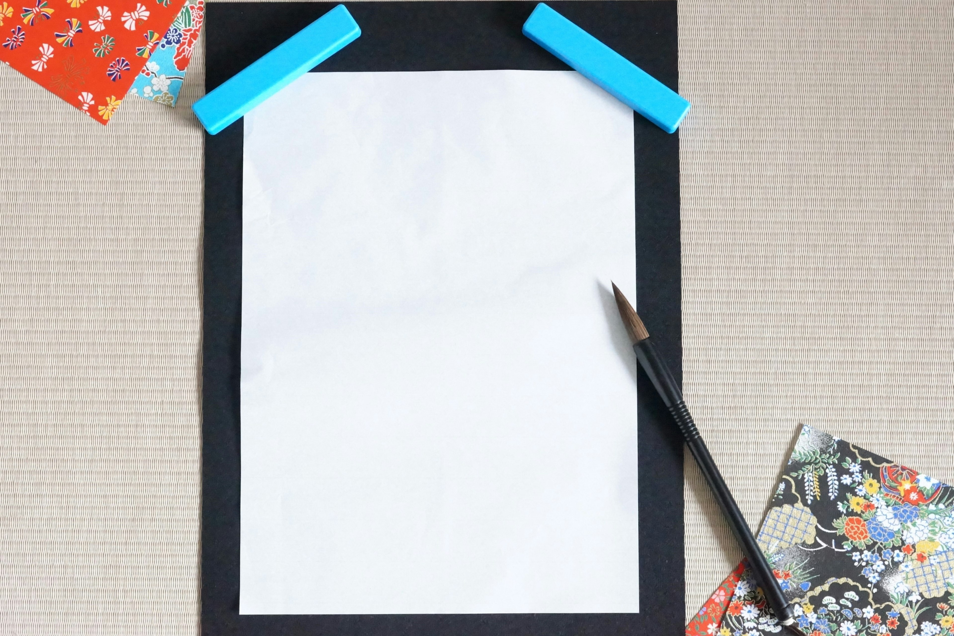 Blank white paper in a black frame with blue clips and a pencil surrounded by colorful patterned papers