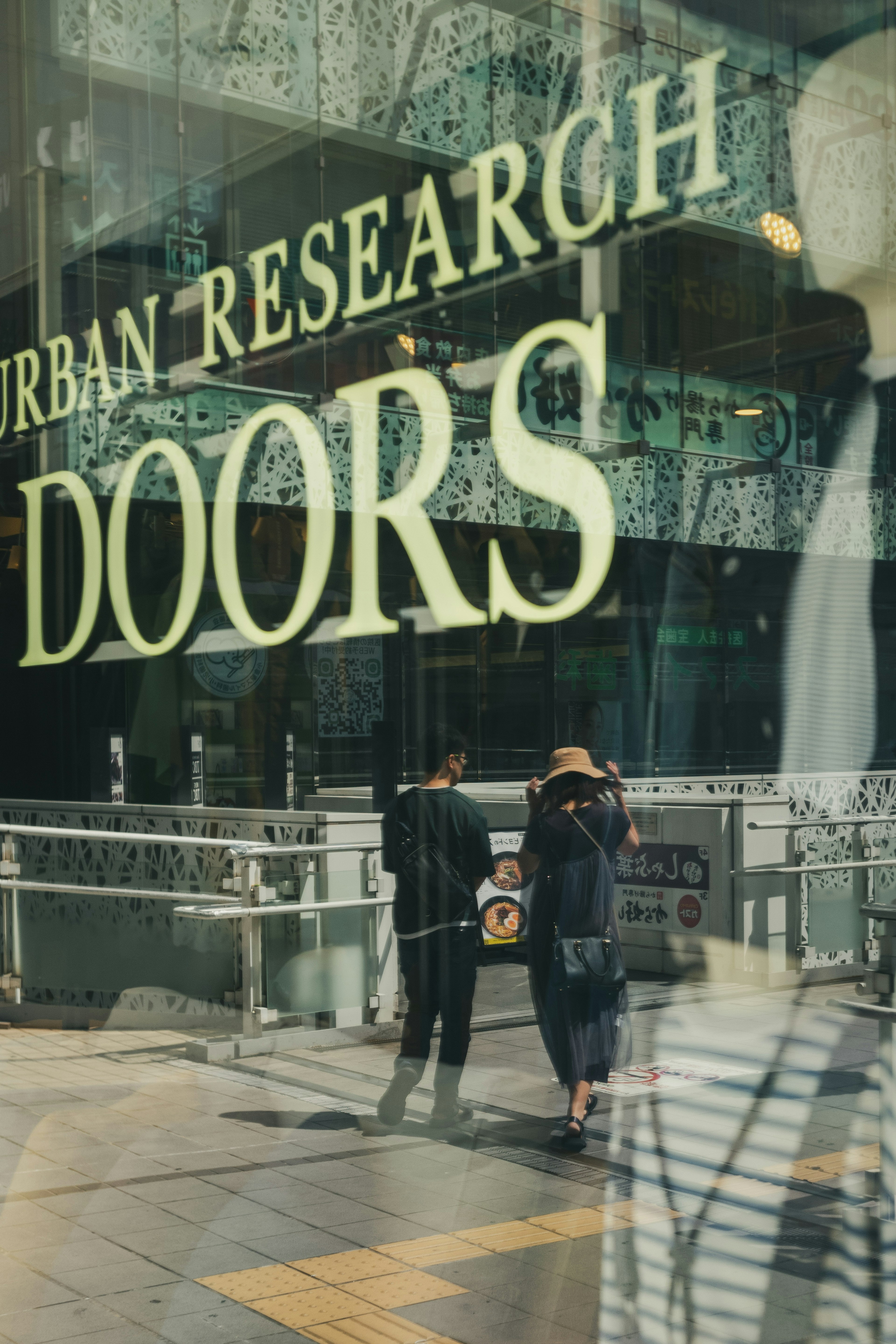 Réflexion de deux personnes marchant devant les portes d'Urban Research