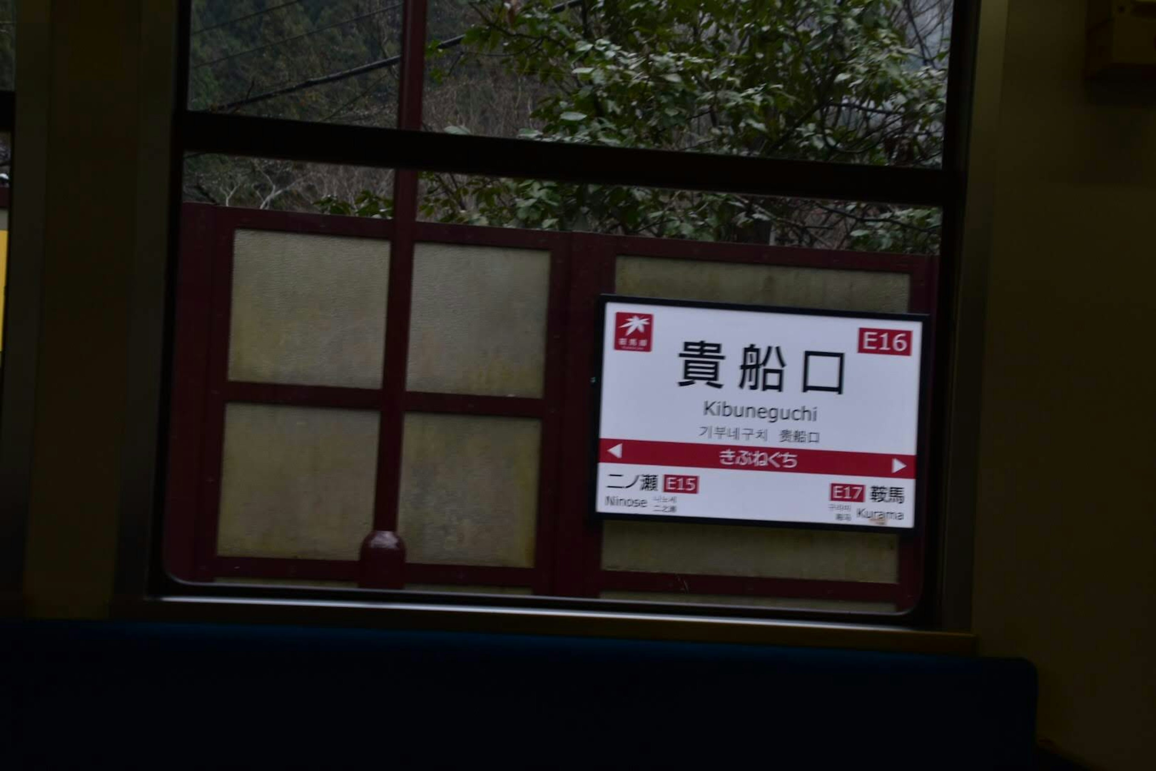 Bahnhofsschild sichtbar aus dem Fenster, weißes und rotes Design, Kanji-Zeichen, umgeben von grünem Gelände