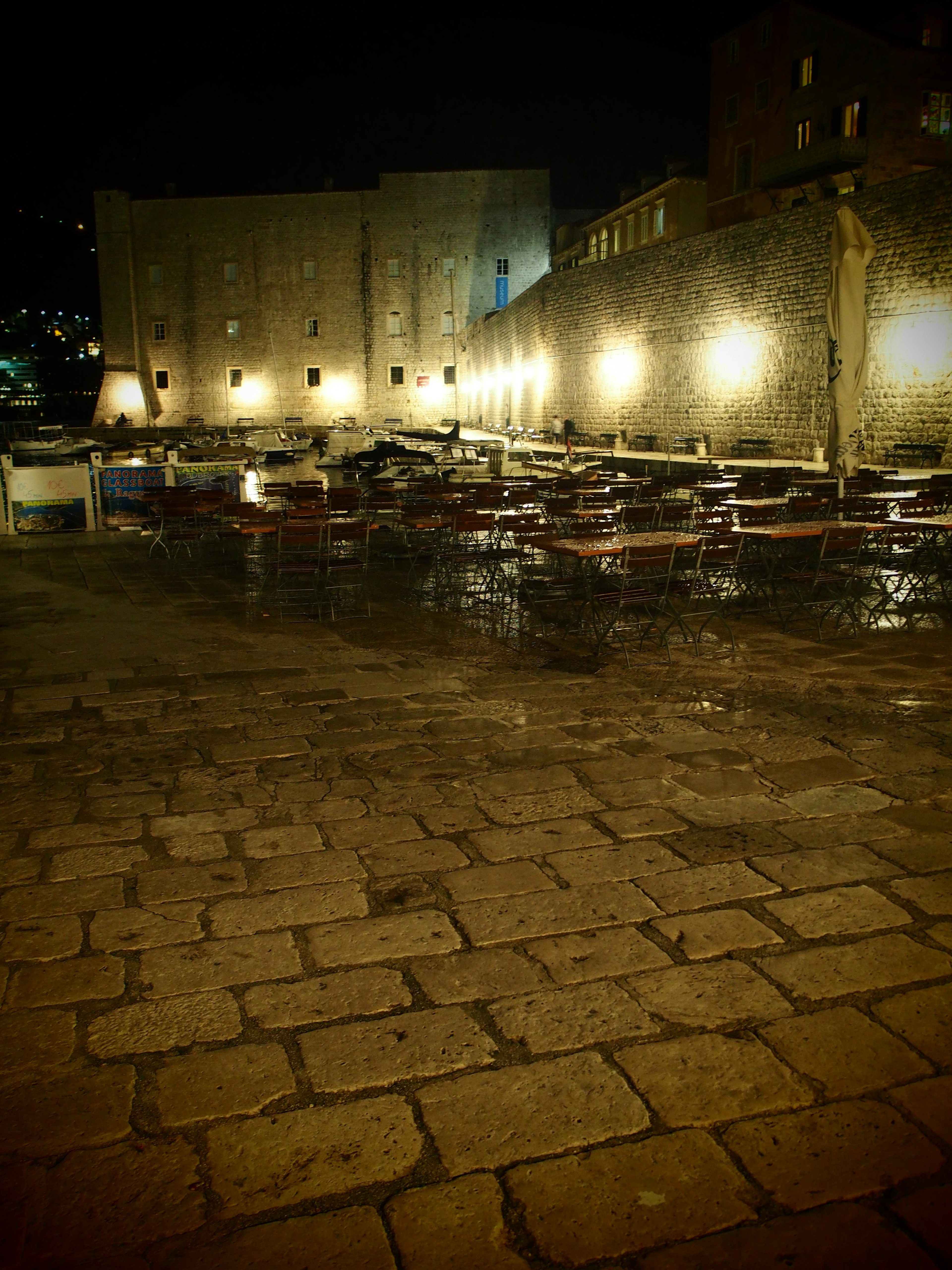 Scène nocturne d'un port avec des reflets de lumières sur des pavés humides et des tables