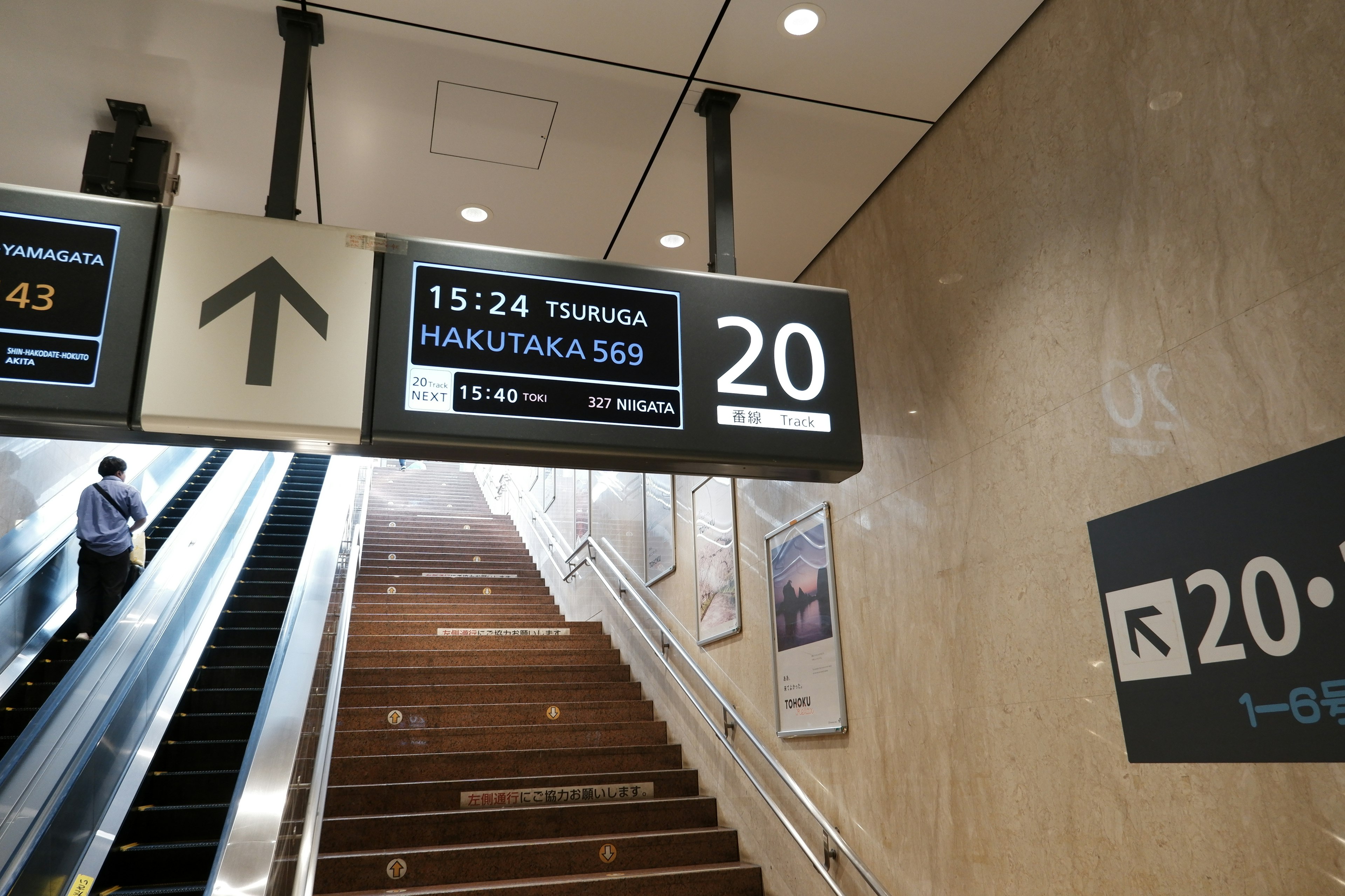 エスカレーターの上にある駅の表示と時刻表が見える