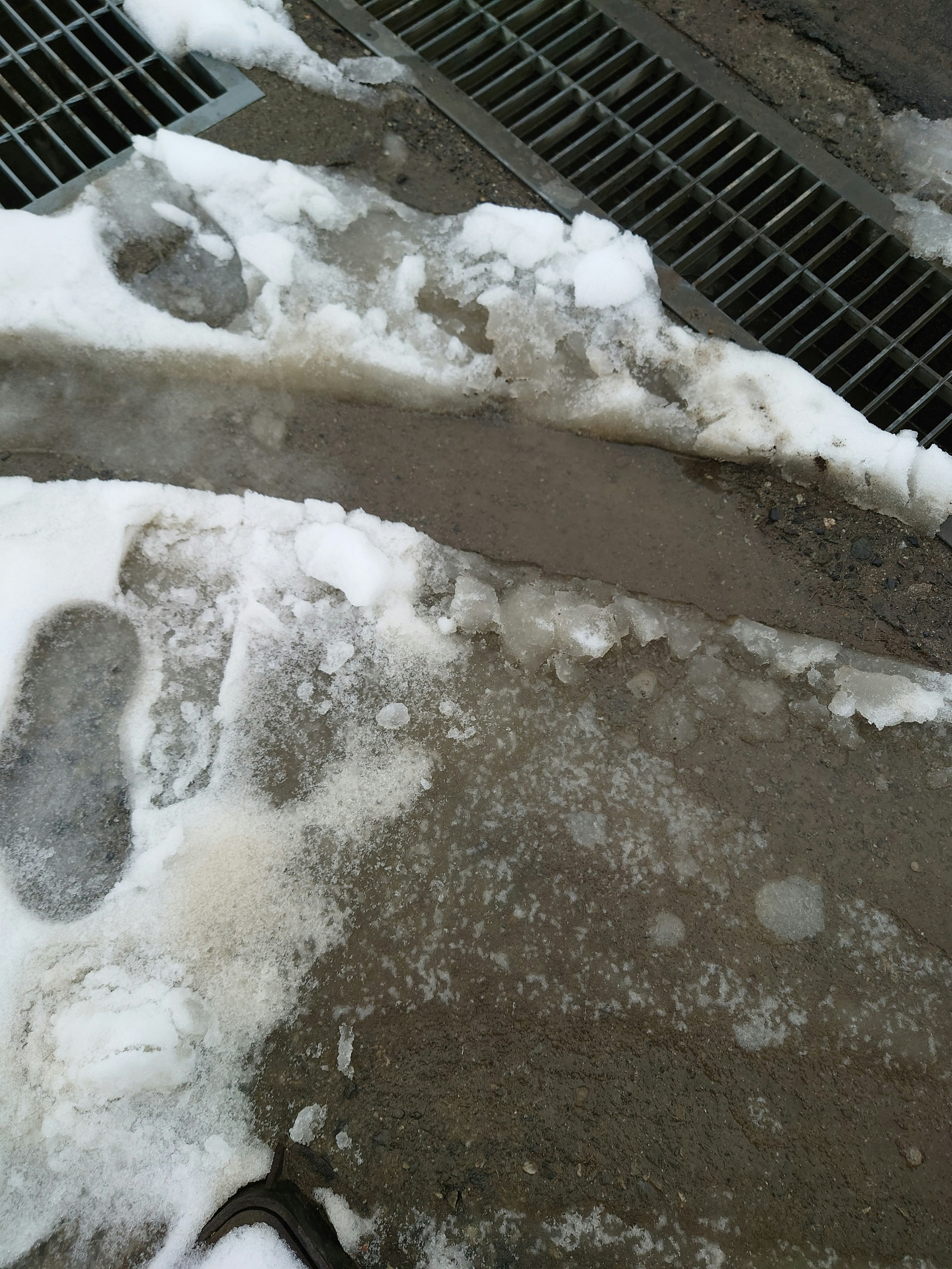 Part of a sidewalk with remaining snow and ice Melted snow and puddles Visible grate