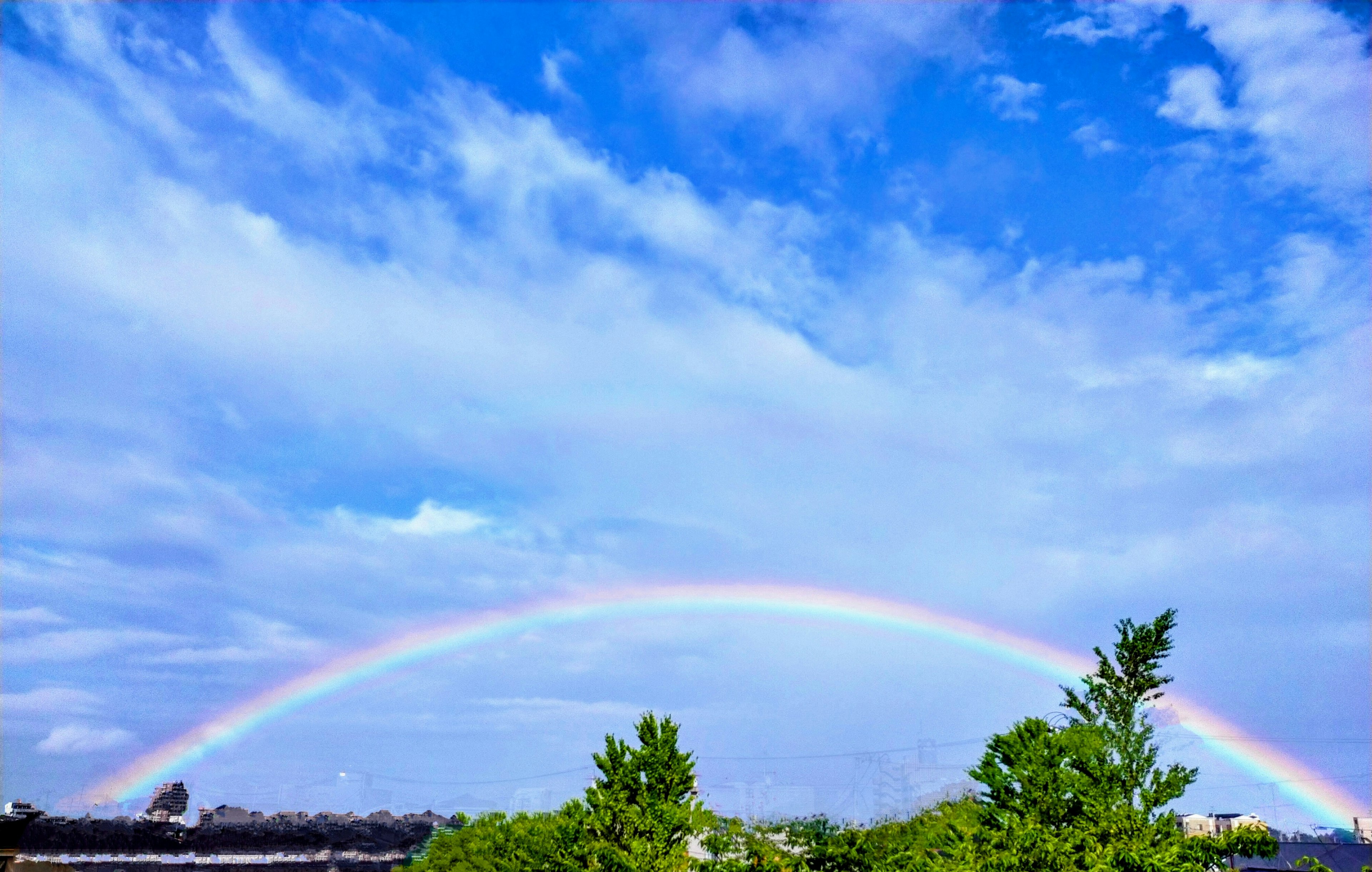 A vibrant rainbow arching across a blue sky with scattered clouds and green trees