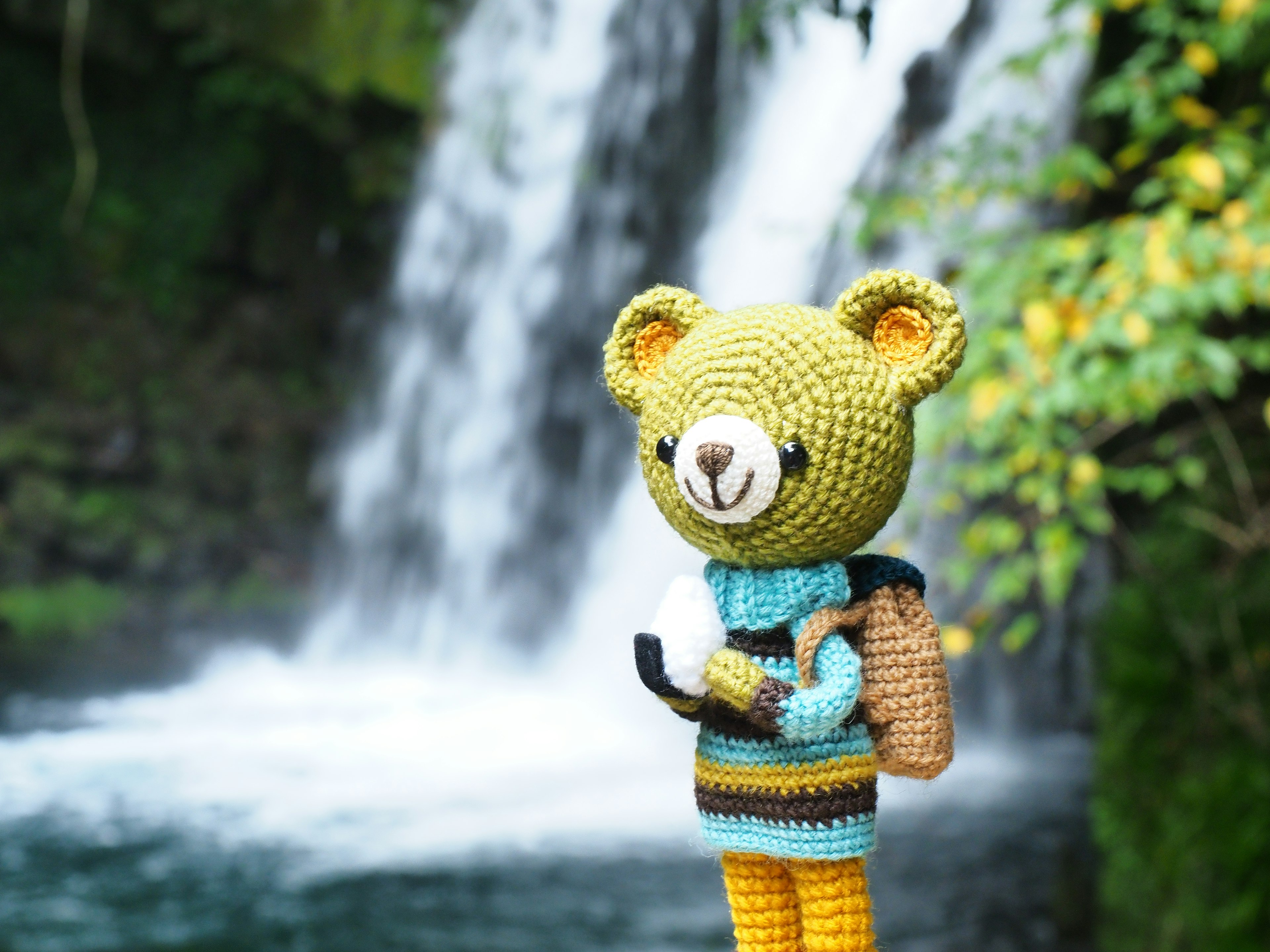 Crocheted bear character standing in front of a waterfall holding a phone