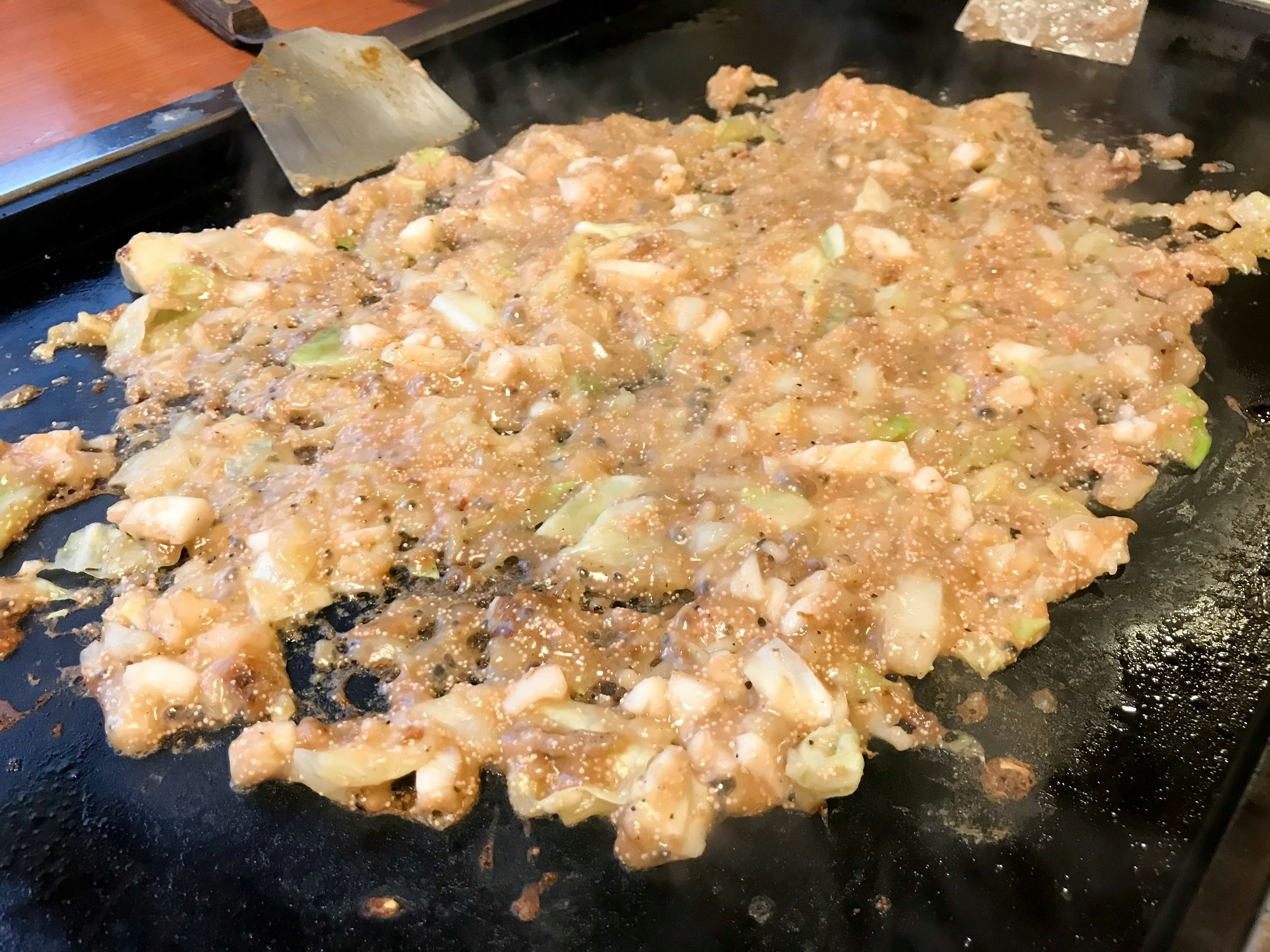 Phần okonomiyaki đang nấu trên vỉ nướng với các nguyên liệu trộn lẫn