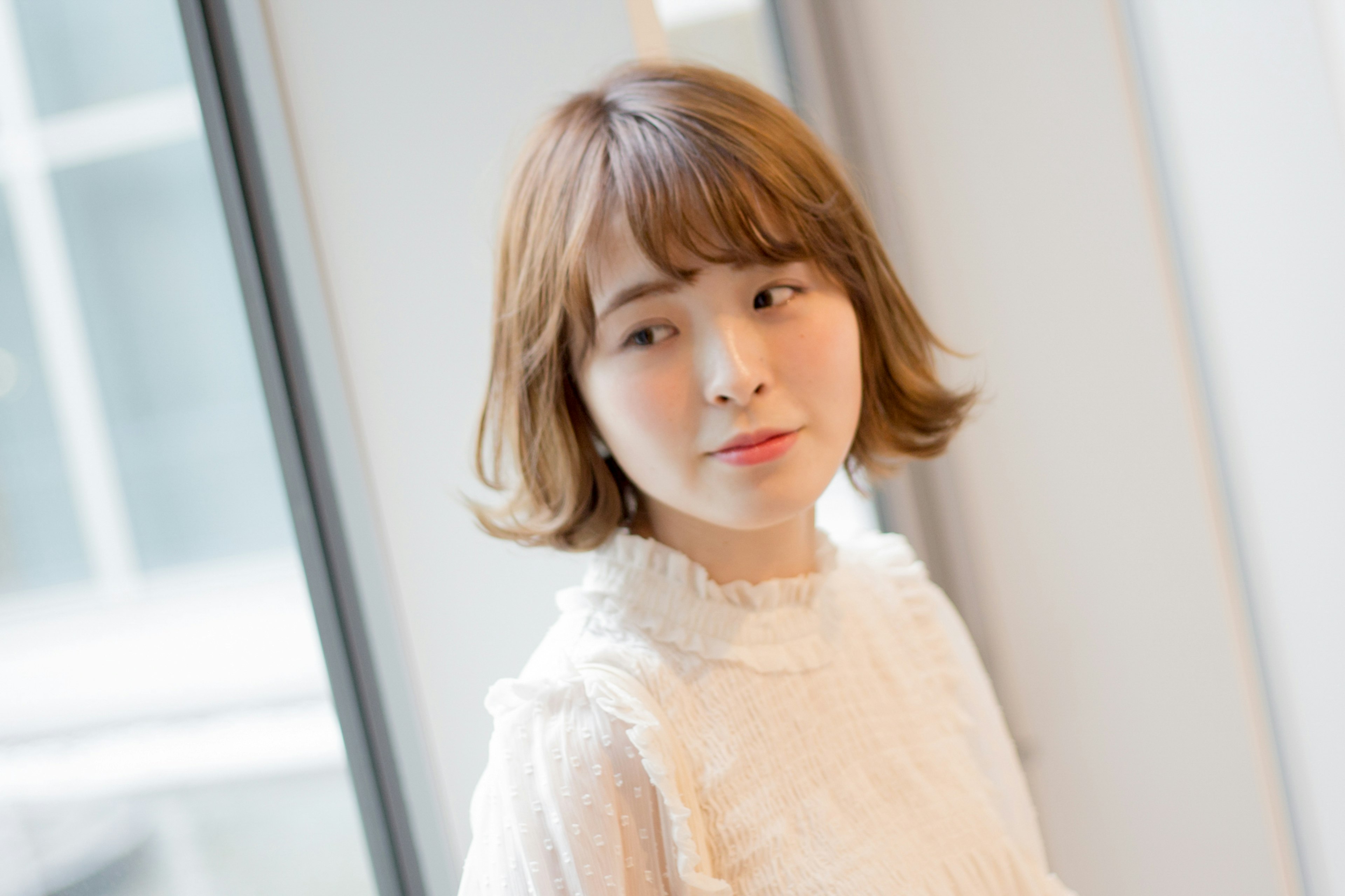 A young woman with short brown hair wearing a white blouse standing near a window