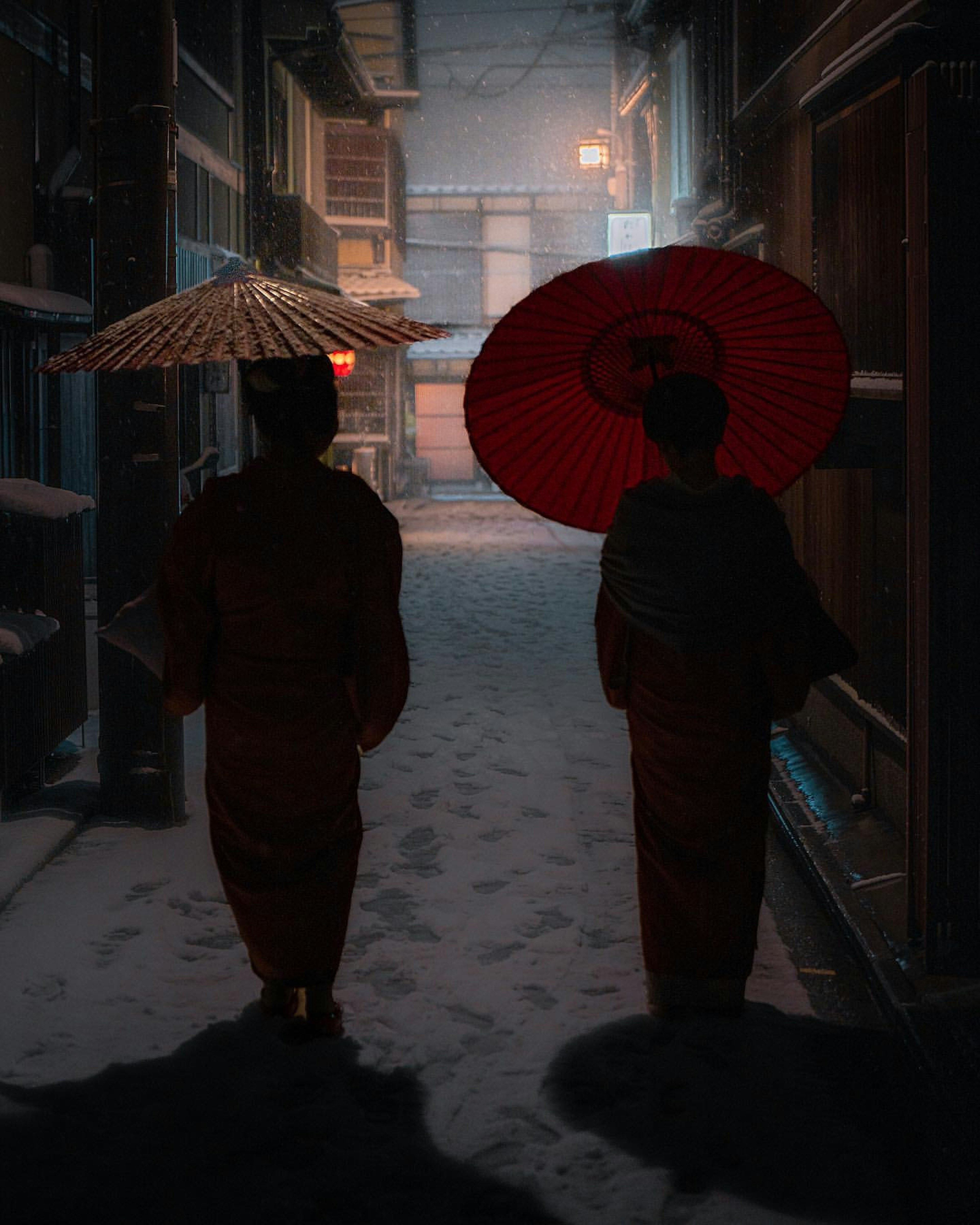 Two figures in kimonos walking under umbrellas in a snowy alleyway