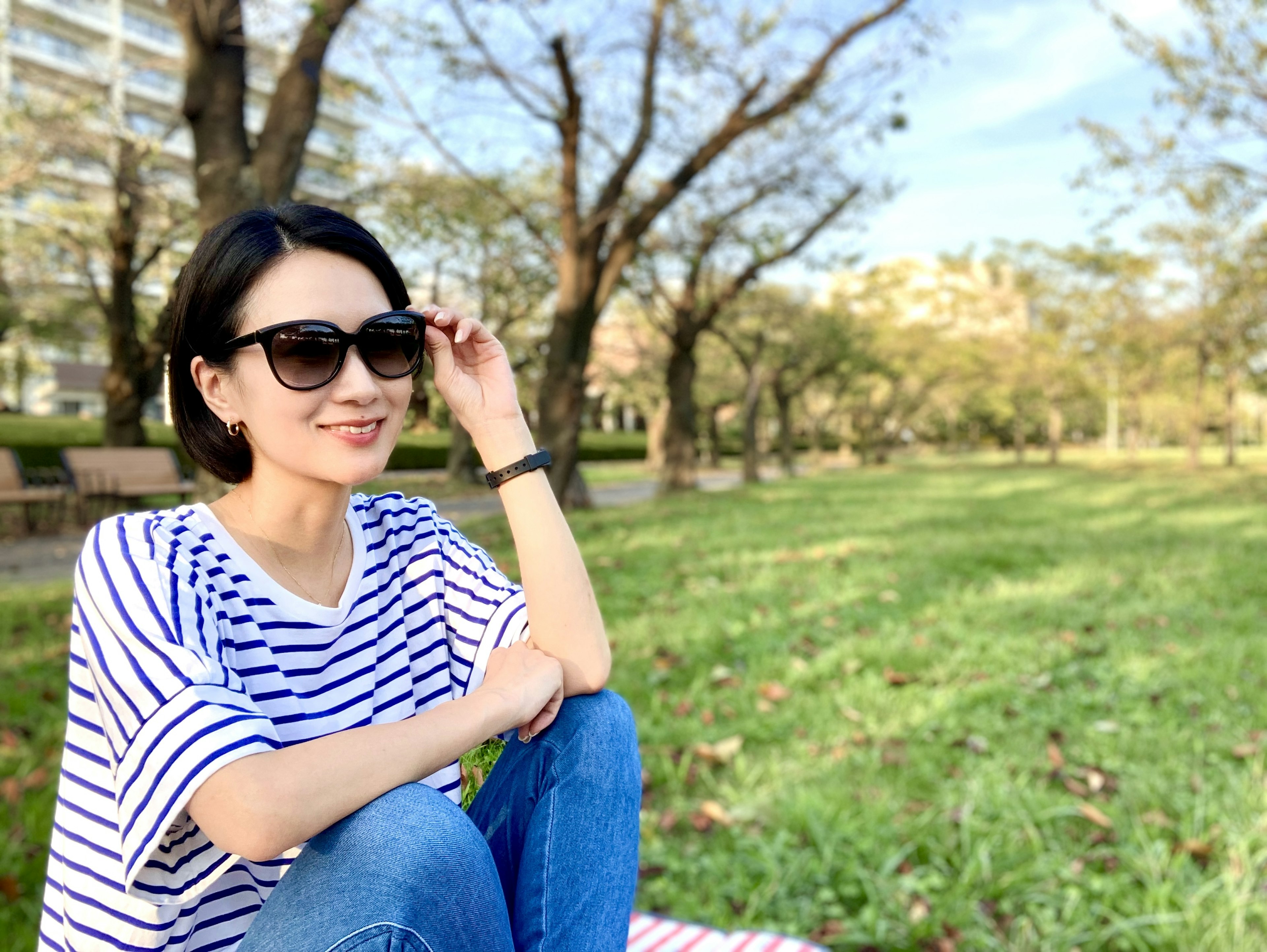 A woman wearing sunglasses sitting in a park with trees and grass