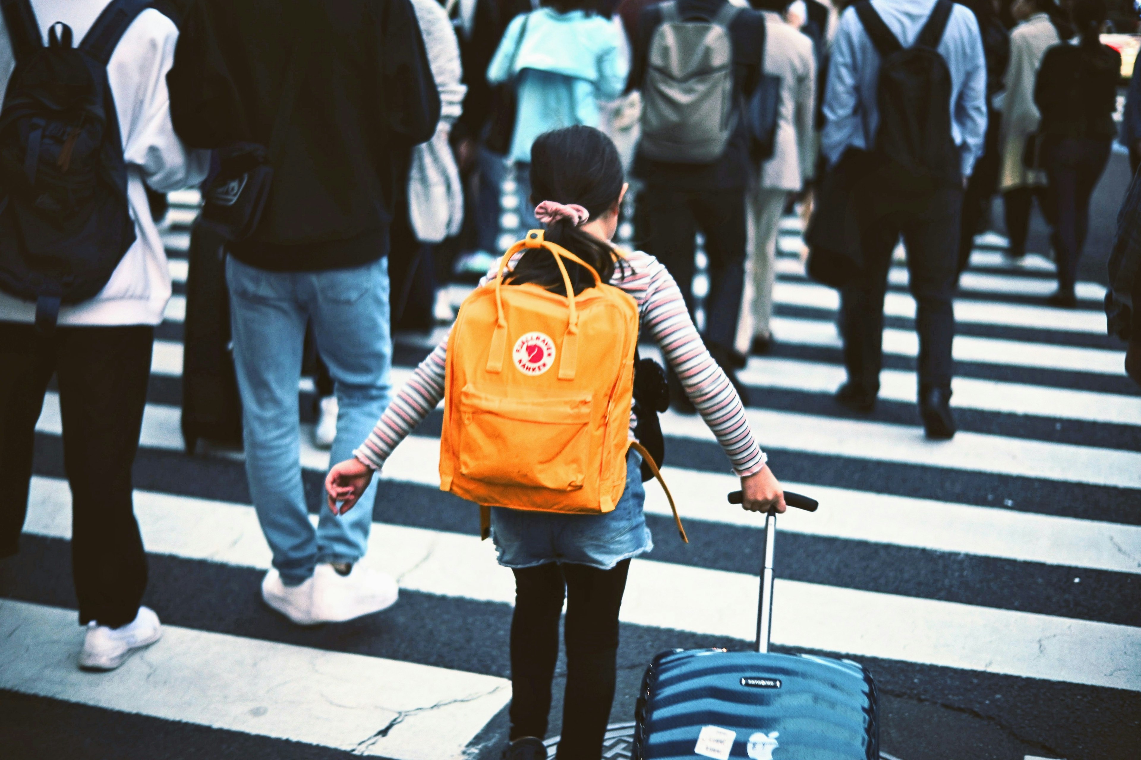 Un niño cruzando la calle con una maleta y una mochila naranja