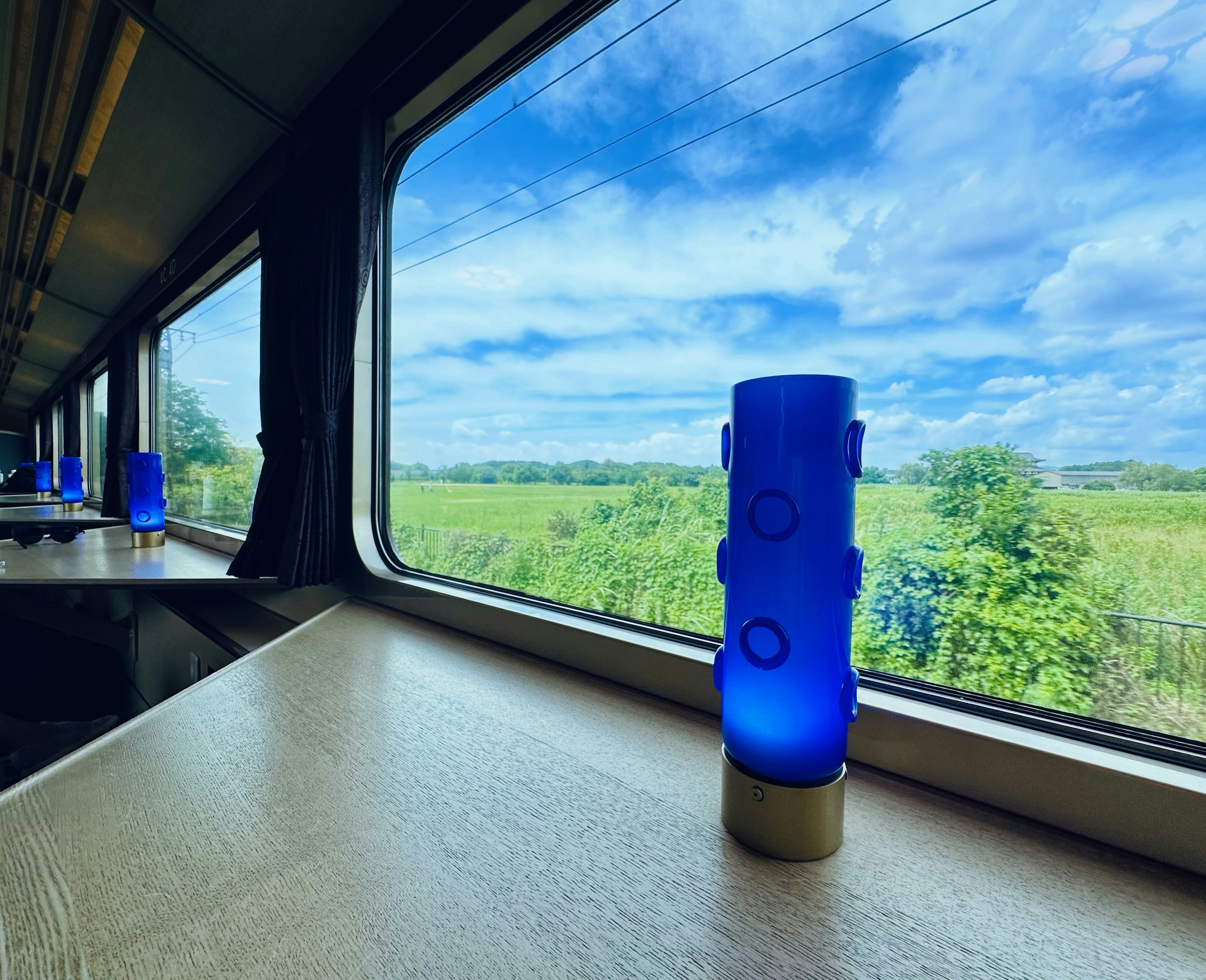 Vaso de vidrio azul en una mesa de tren con paisaje verde y cielo azul visible por la ventana