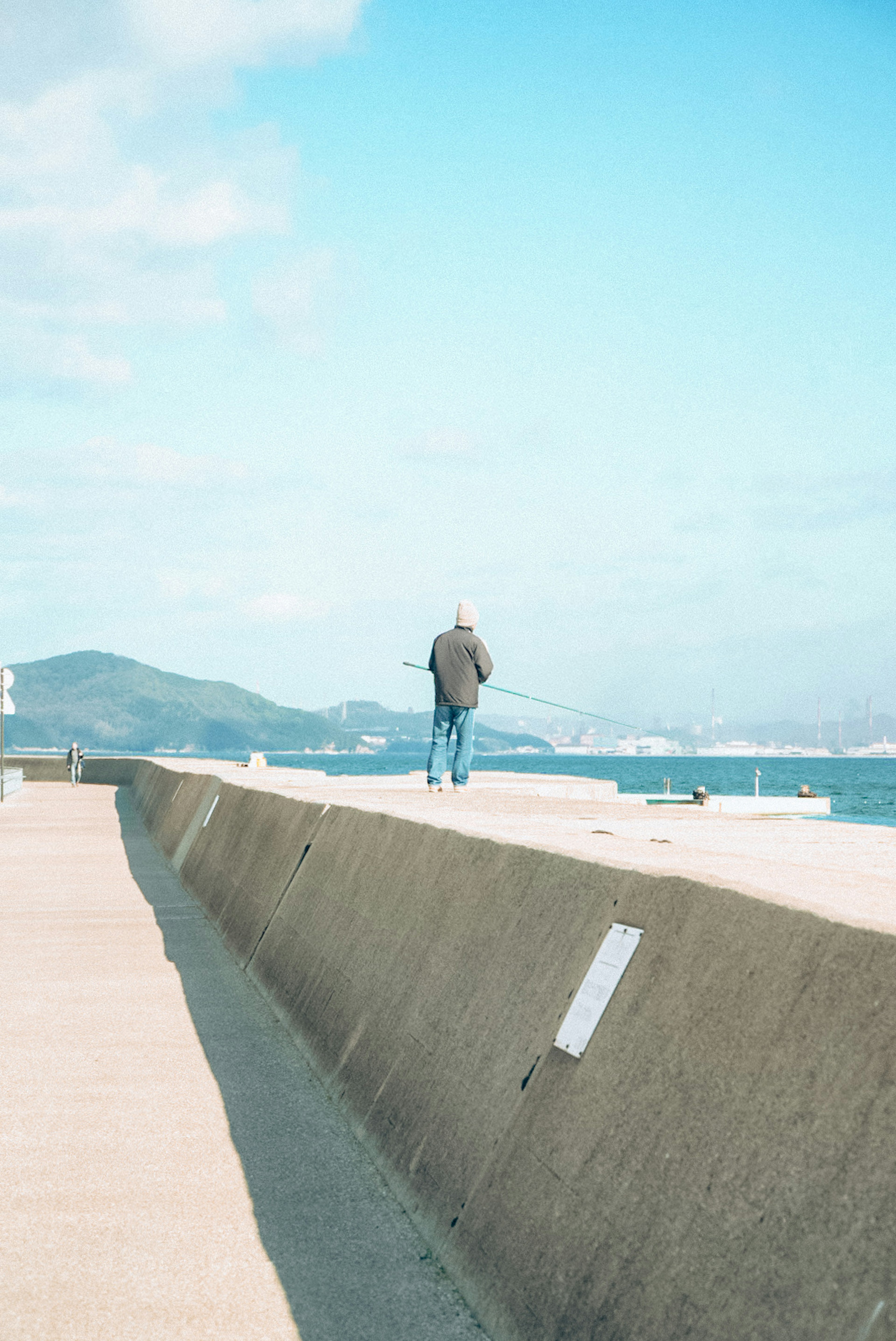 Una persona caminando por un rompeolas costero con cielo azul y montañas distantes