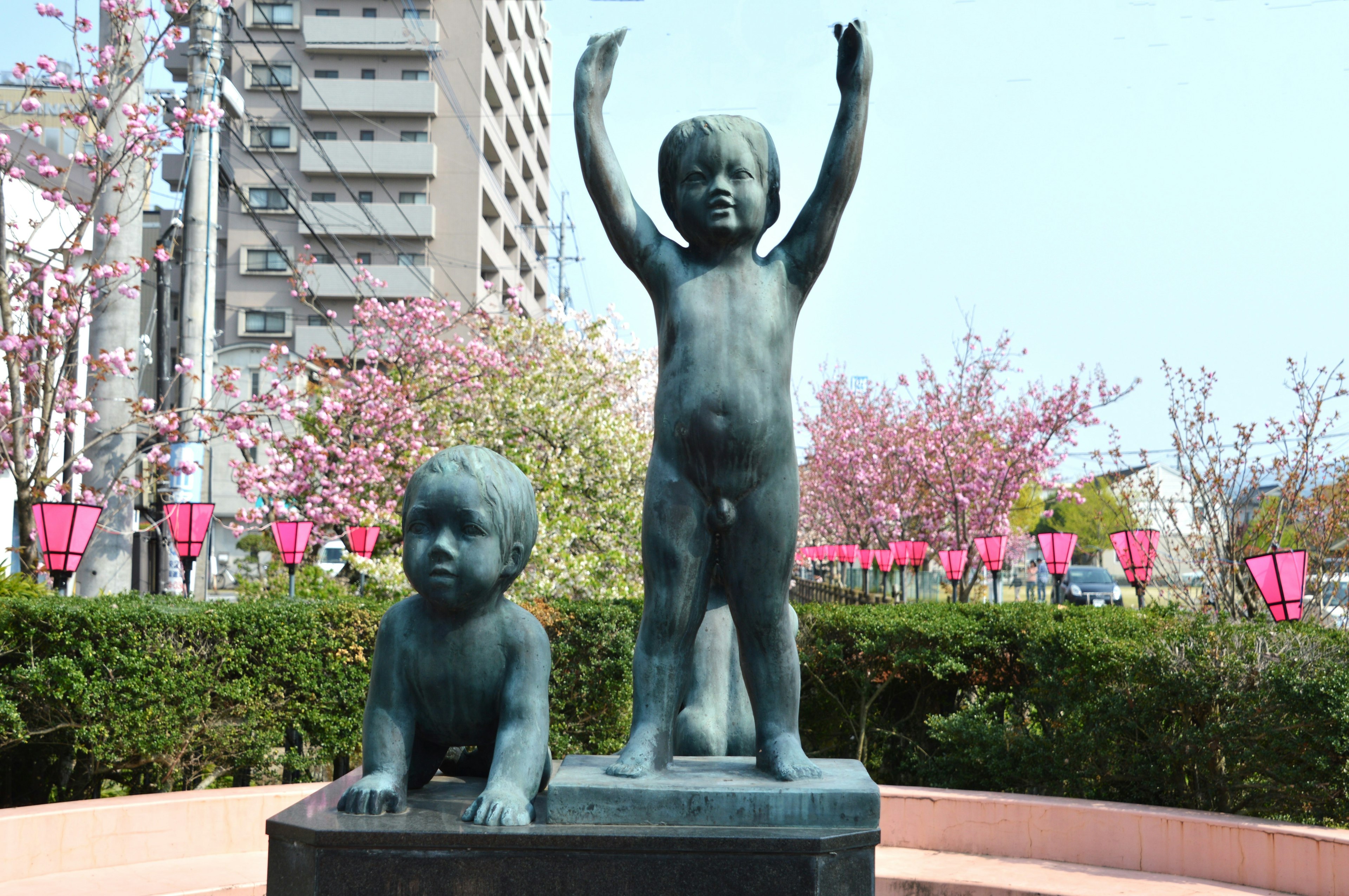 Bronze statue of children in a park, one standing with arms raised, the other crawling, with cherry blossom trees and buildings in the background