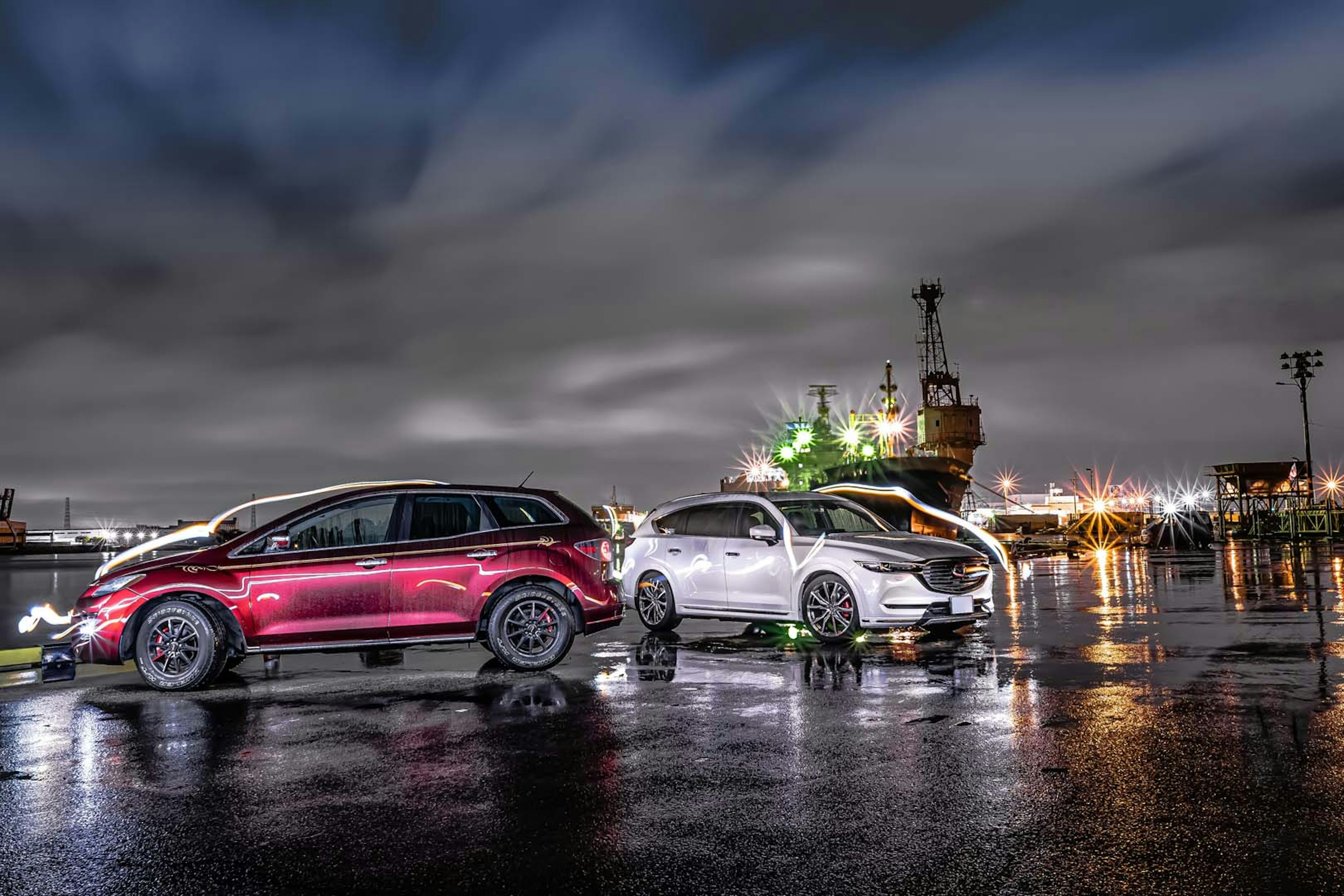 Red and silver cars parked at night in a harbor with reflections on wet pavement