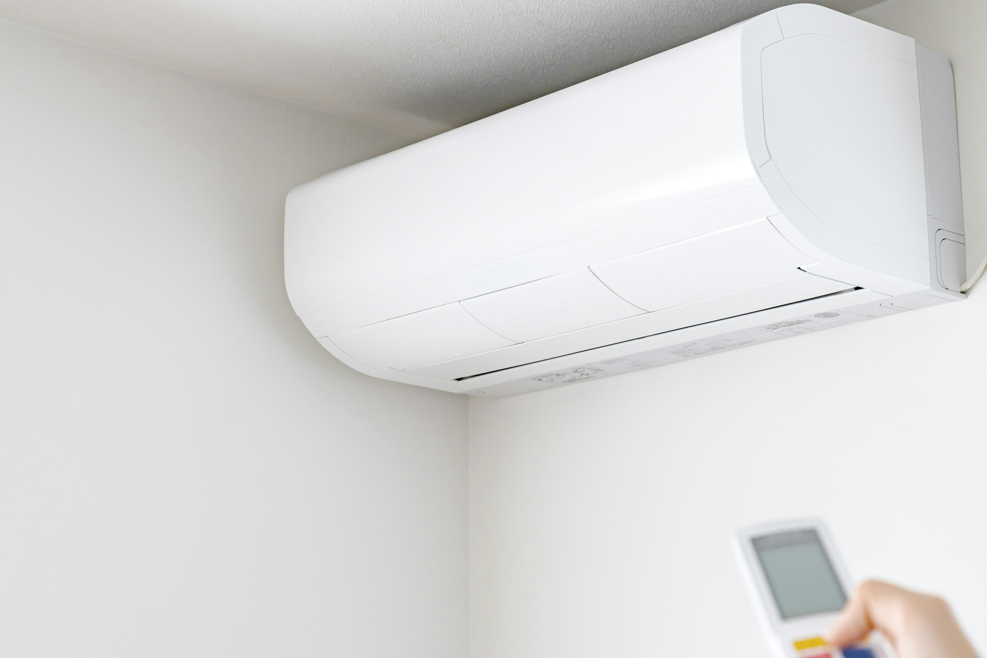 A white air conditioner mounted on the wall with a hand holding a remote control