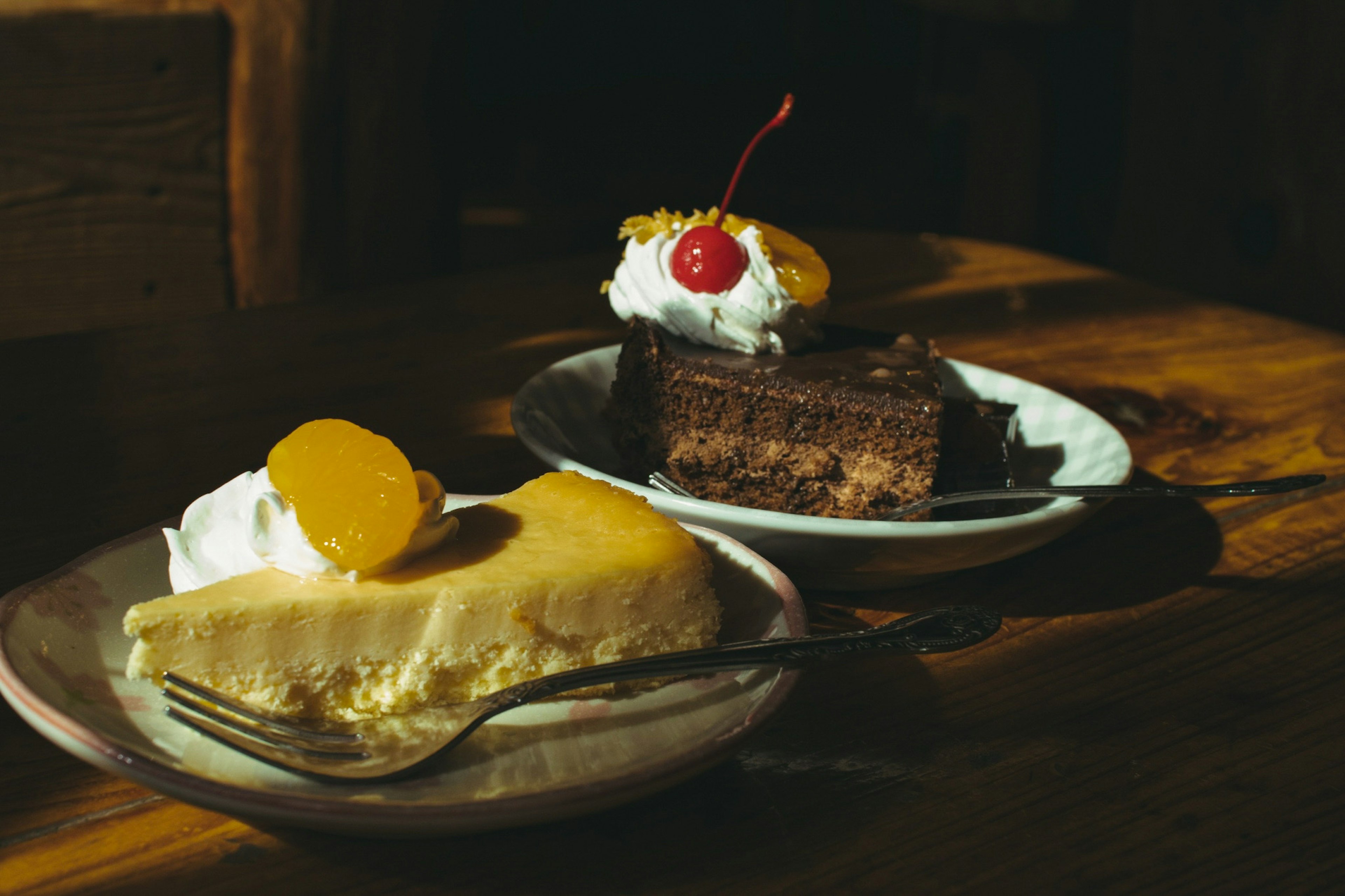 Pastel de chocolate cubierto con crema y cereza junto a un cheesecake con mango