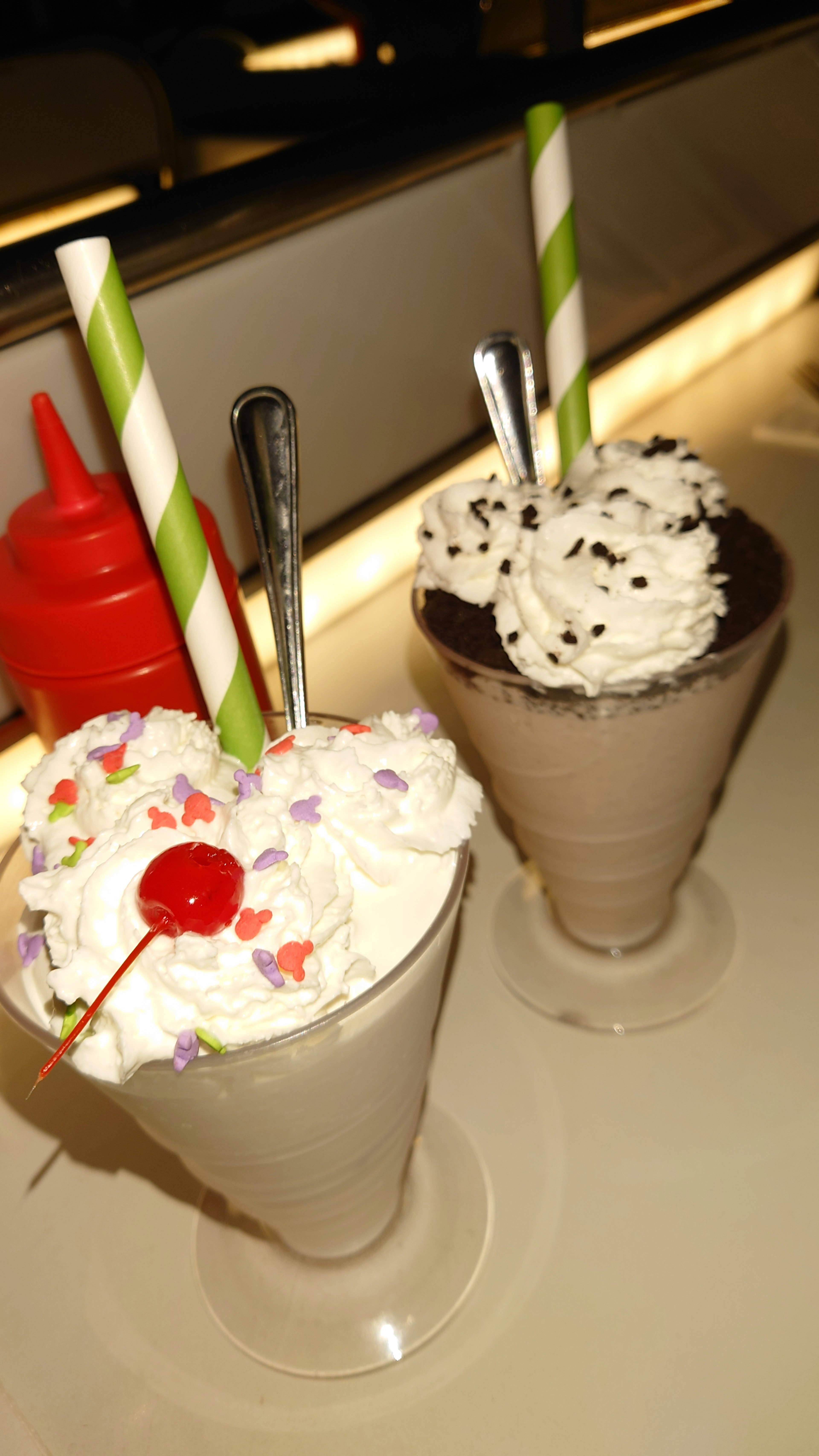 Dua milkshake es krim disajikan dalam gelas dengan krim kocok dan ceri di atas salah satunya dihiasi dengan taburan berwarna