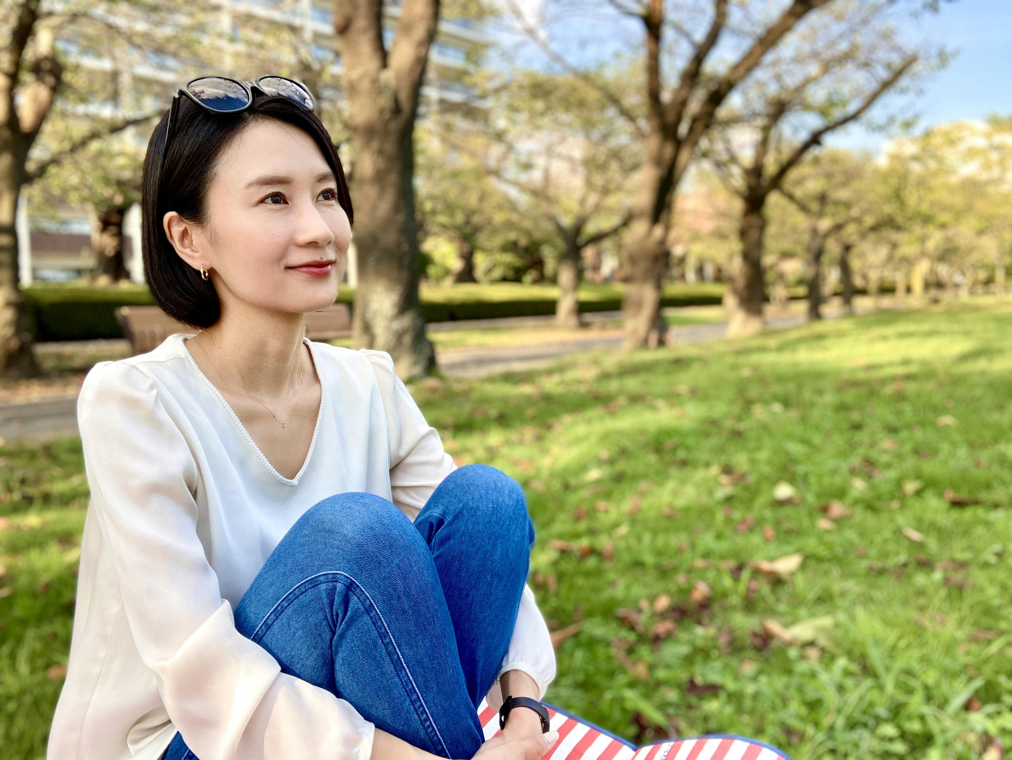 Potret wanita duduk di rumput hijau di taman mengenakan celana jeans biru dan blouse putih dengan pohon di latar belakang