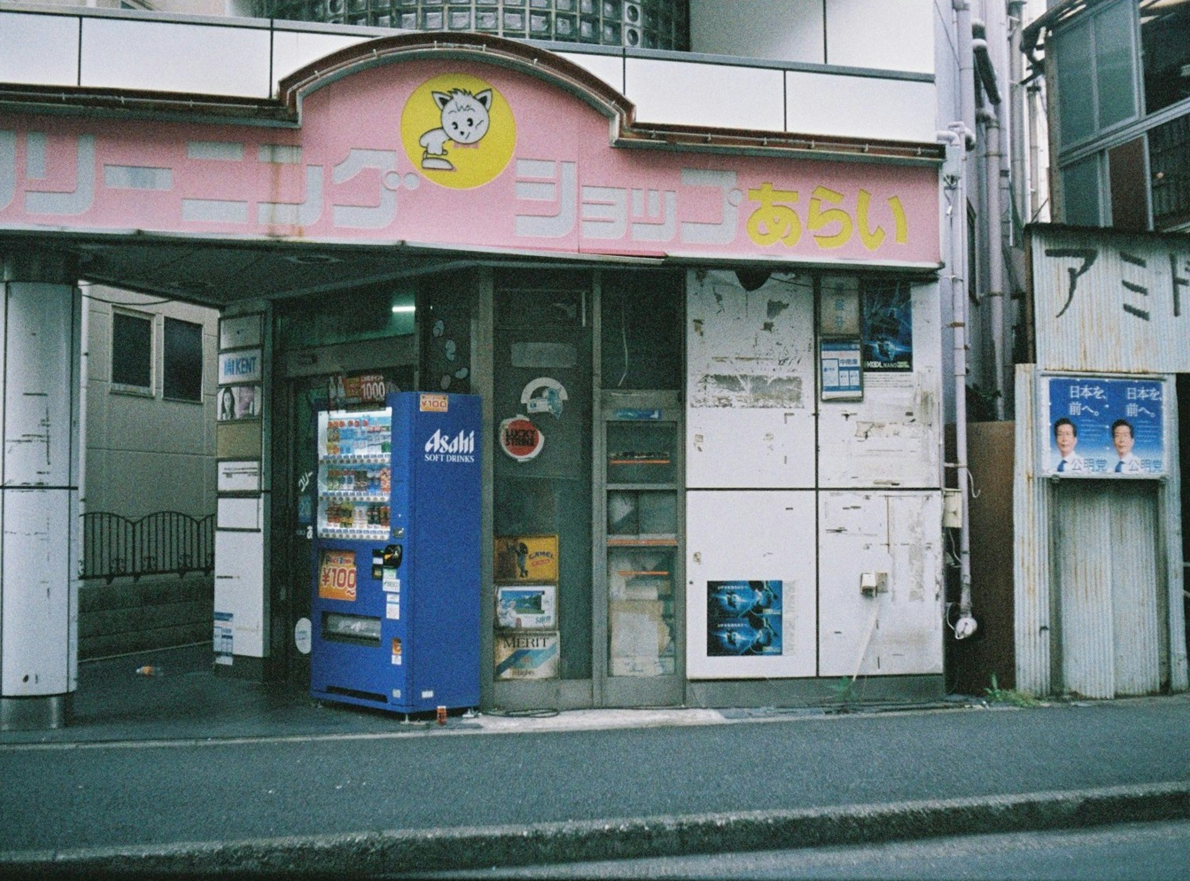 ピンクの看板が特徴的な小さな店舗の入口に自動販売機が置かれている風景