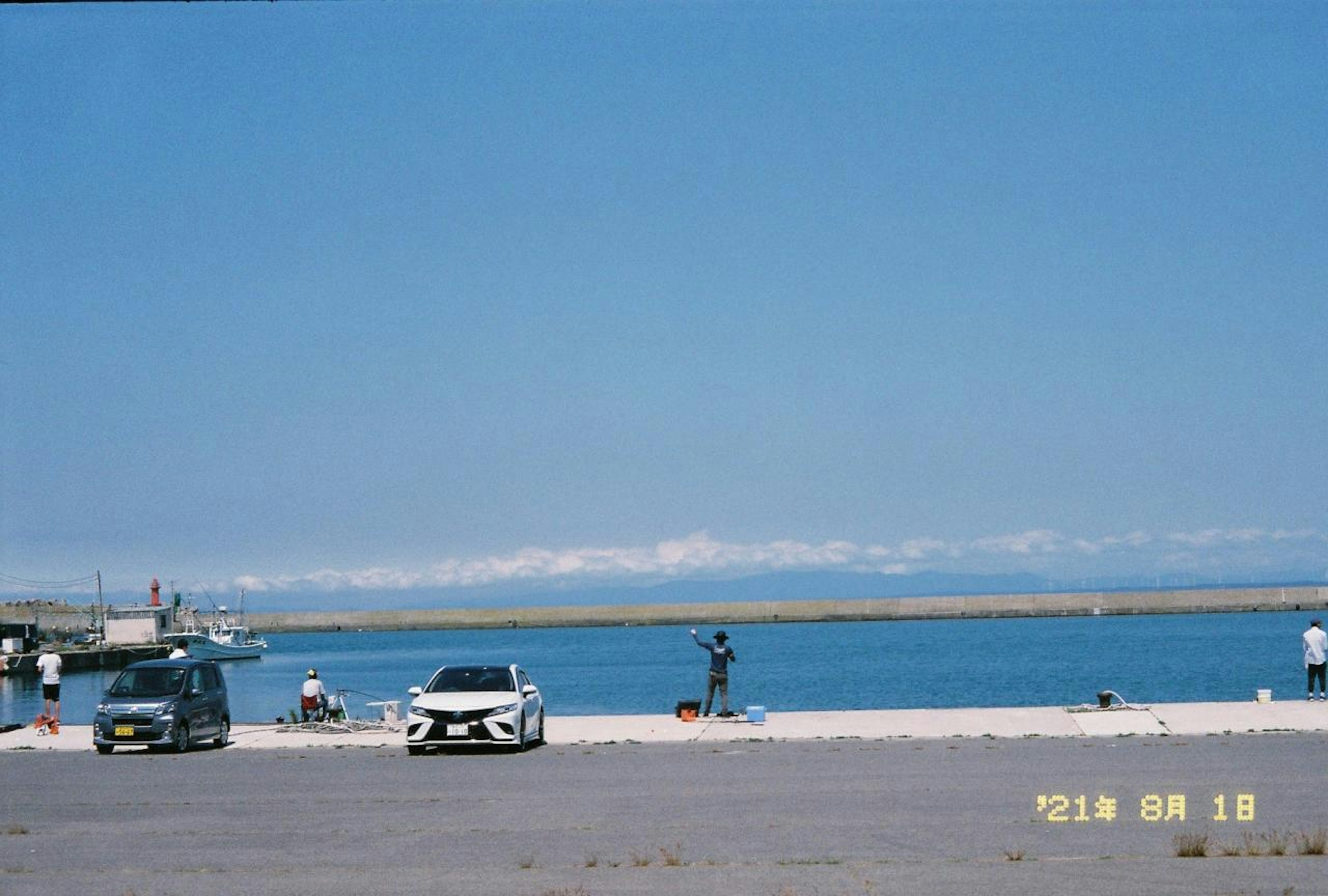 ทิวทัศน์ชายหาดที่มีท้องฟ้าสีฟ้าสดใสและน้ำสงบมีรถจอดและชาวประมง