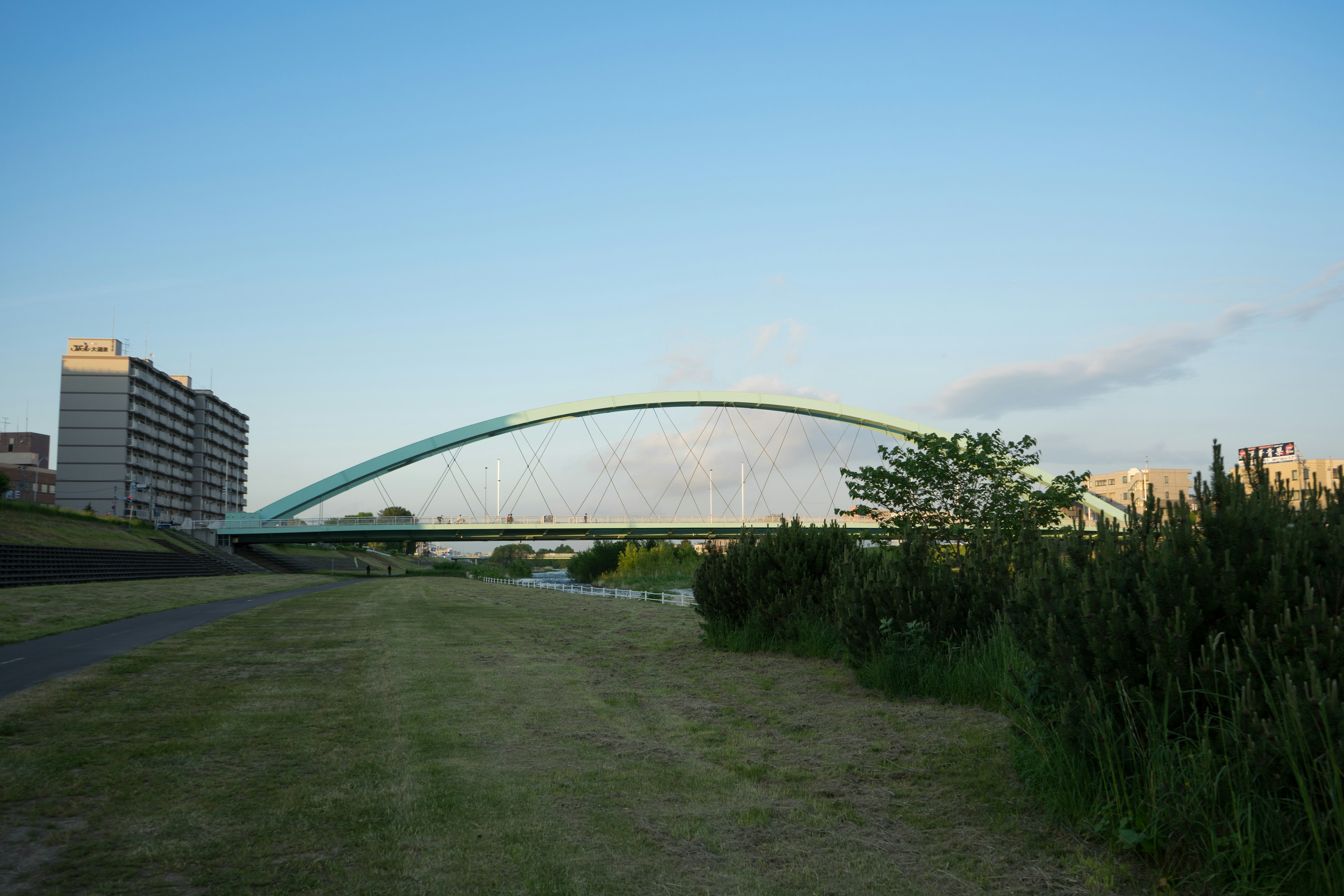 An arch bridge in green spanning over a river with a clear blue sky and grassy area