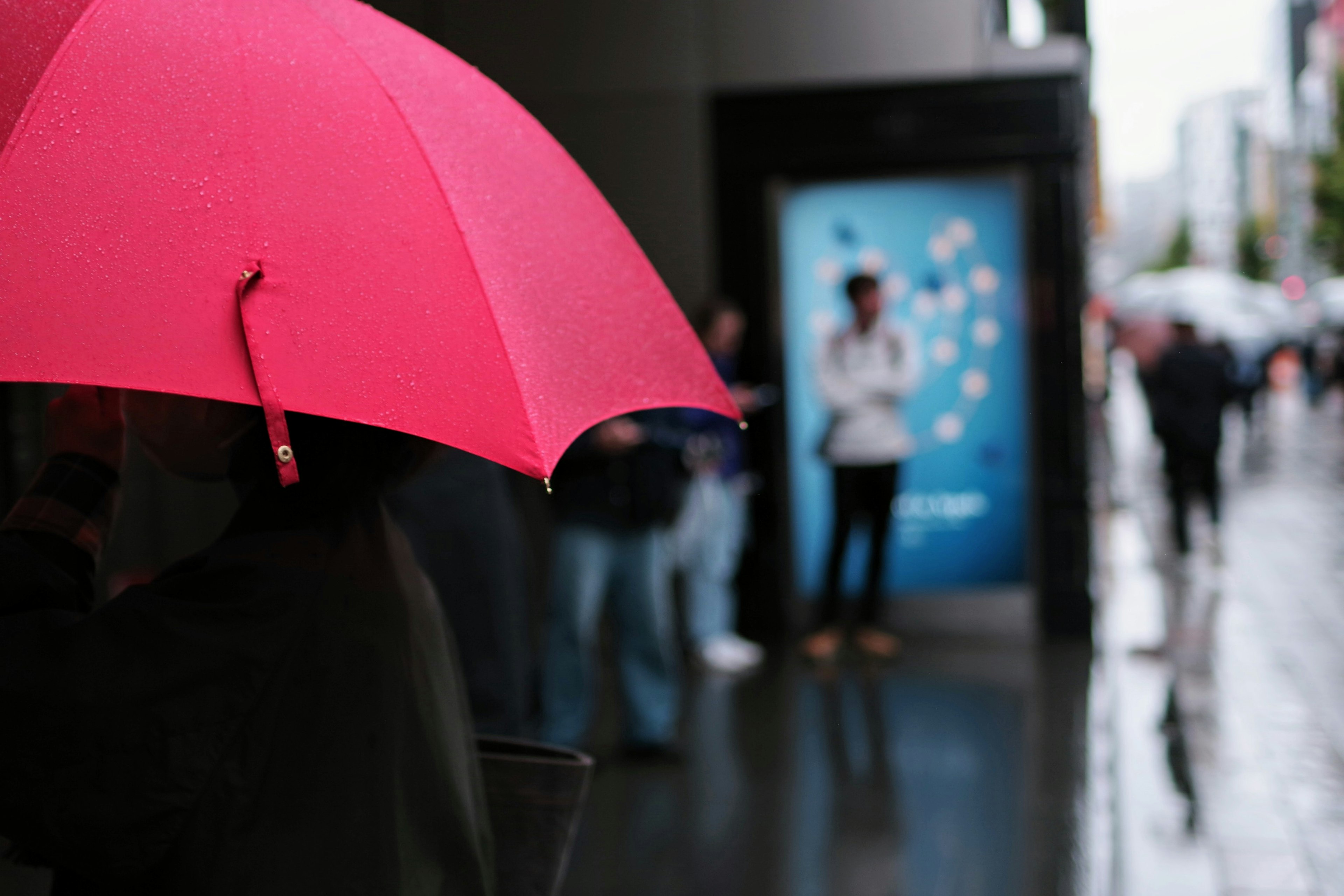 Person holding a pink umbrella in the rain with a blue advertisement in the background