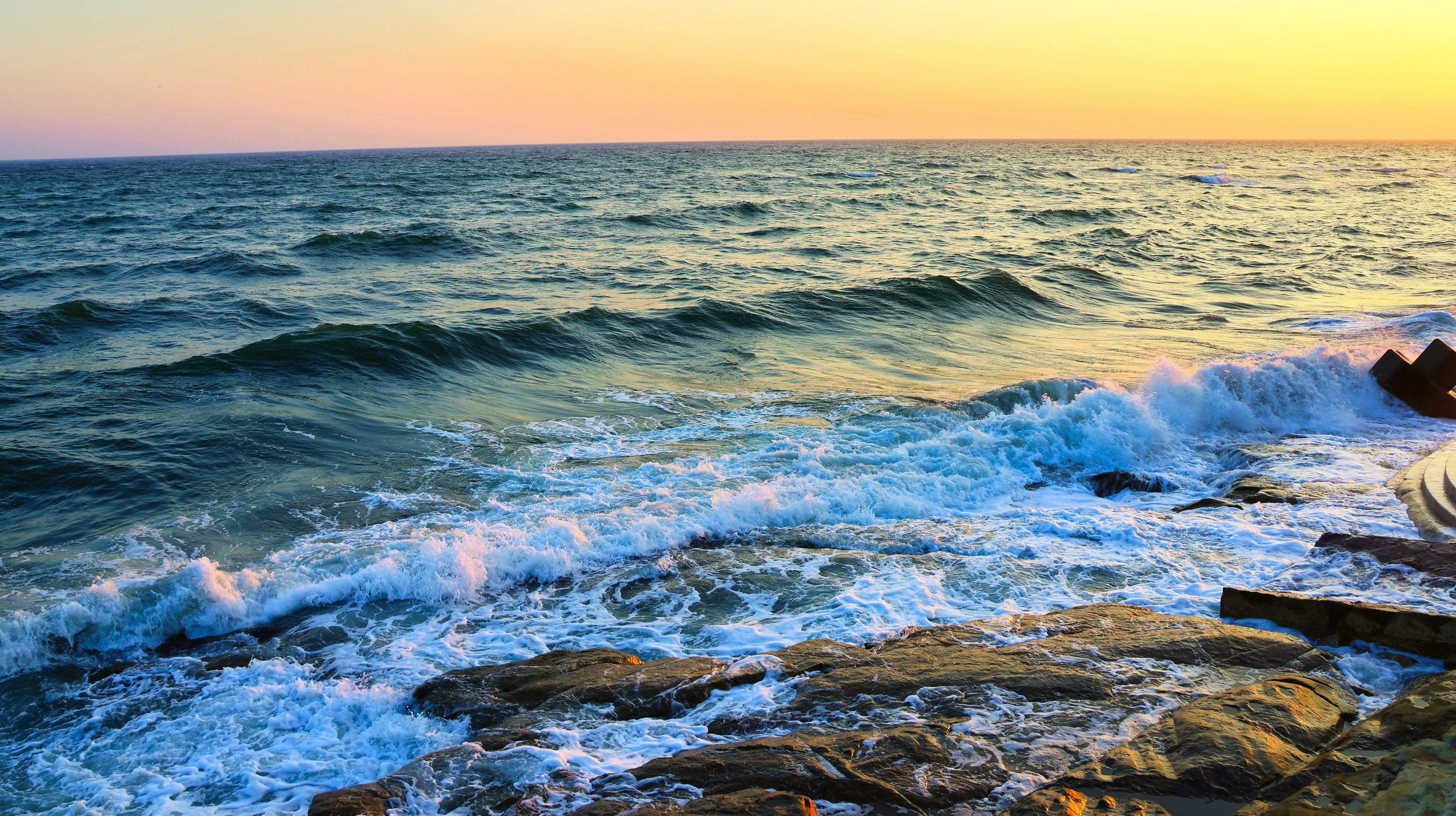 Senja di atas lautan dengan ombak yang menghantam pantai berbatu