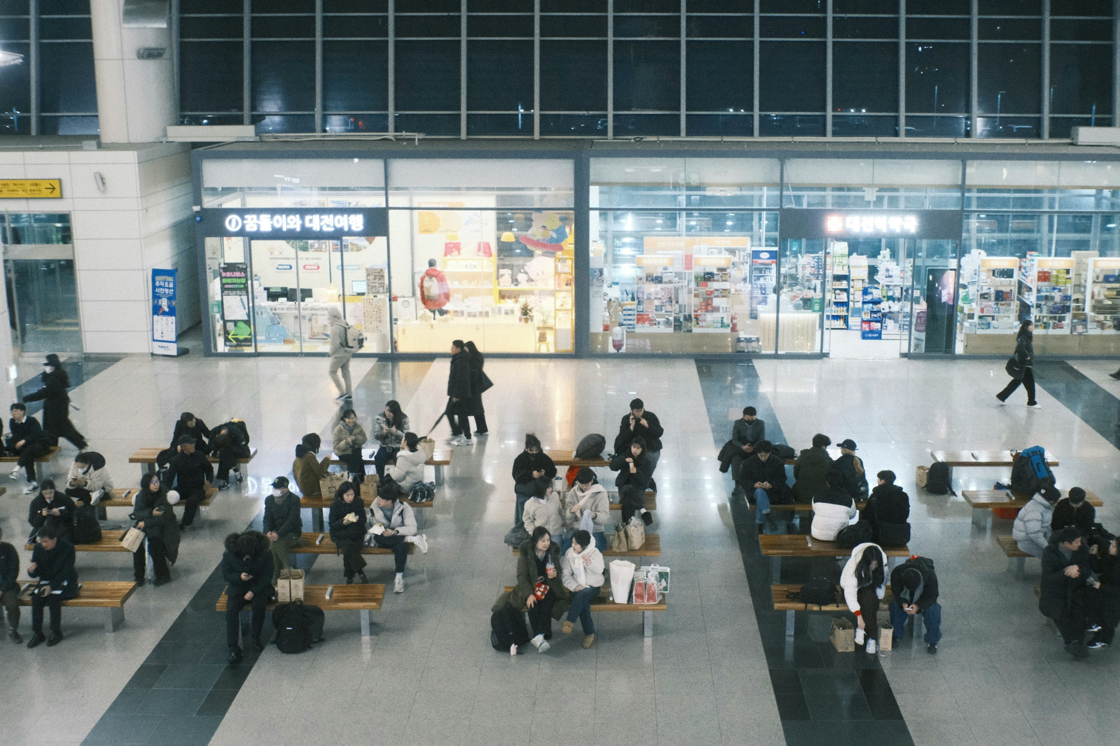 Passagiere in einem geräumigen Warteraum eines Bahnhofs mit Geschäften im Hintergrund