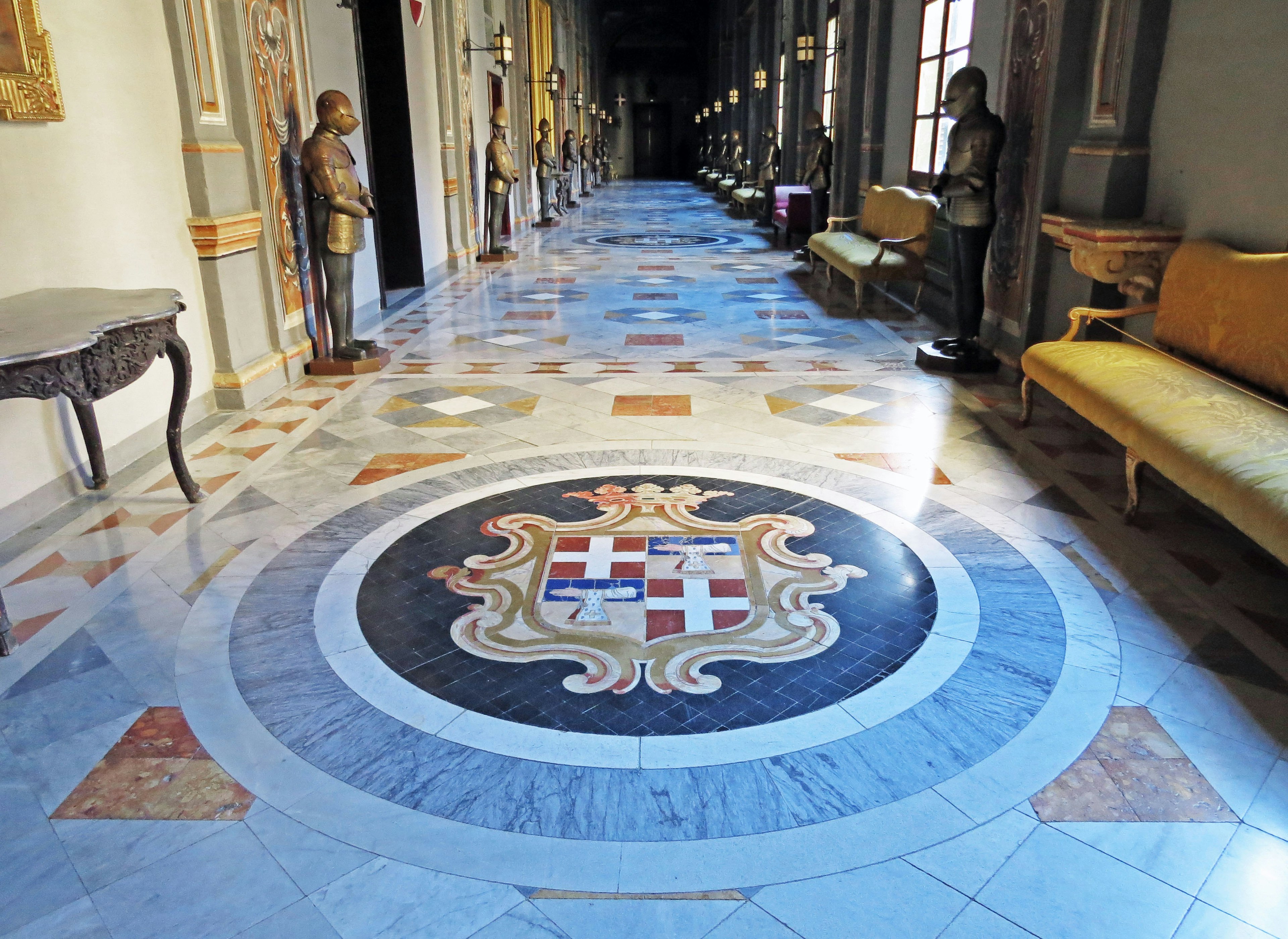 A hallway featuring a beautiful mosaic floor with a central emblem statues and elegant seating along the sides