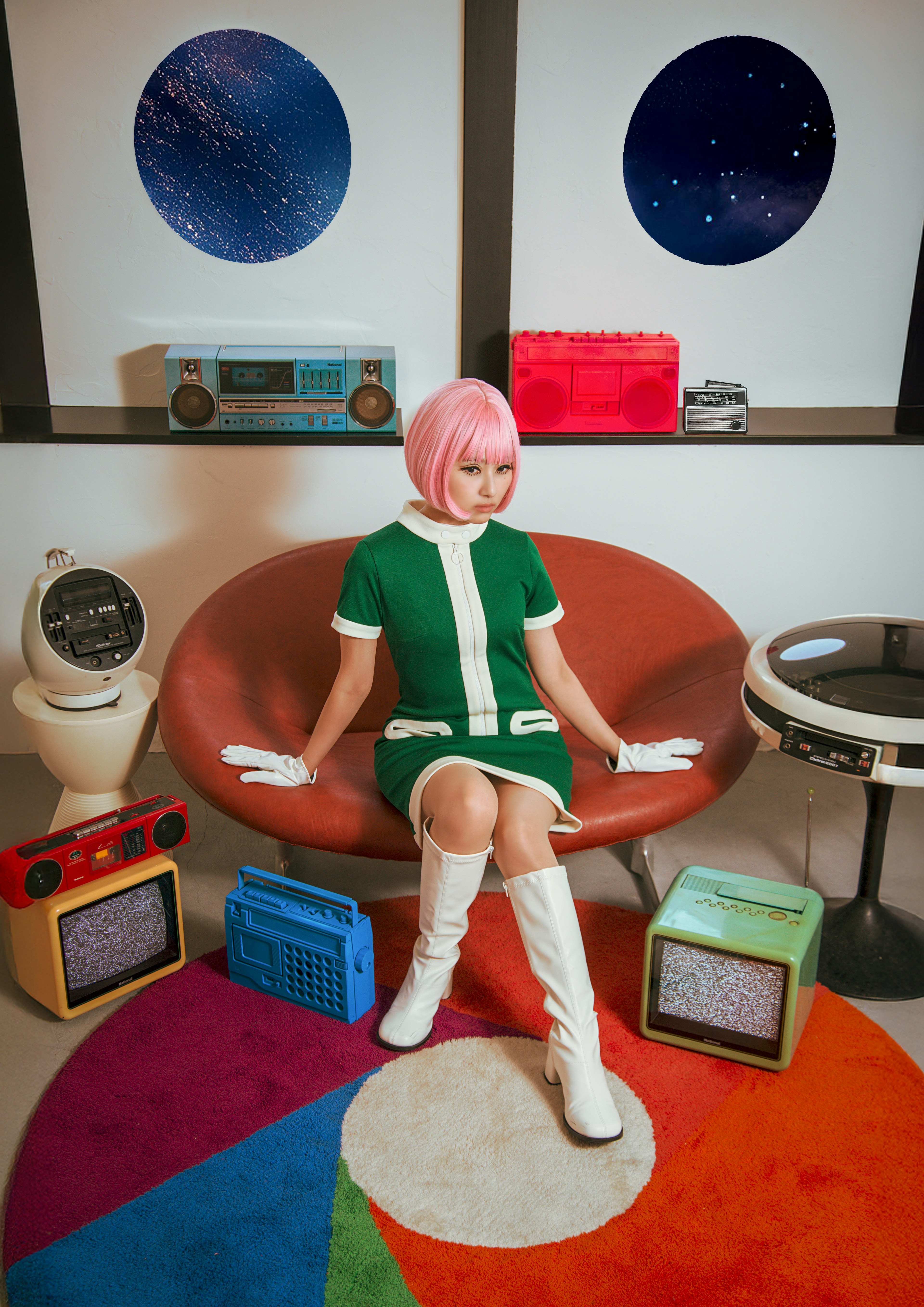 Una mujer con cabello rosa vestida con un vestido verde está sentada en una silla redonda Objetos retro coloridos la rodean incluyendo radios vintage y una alfombra