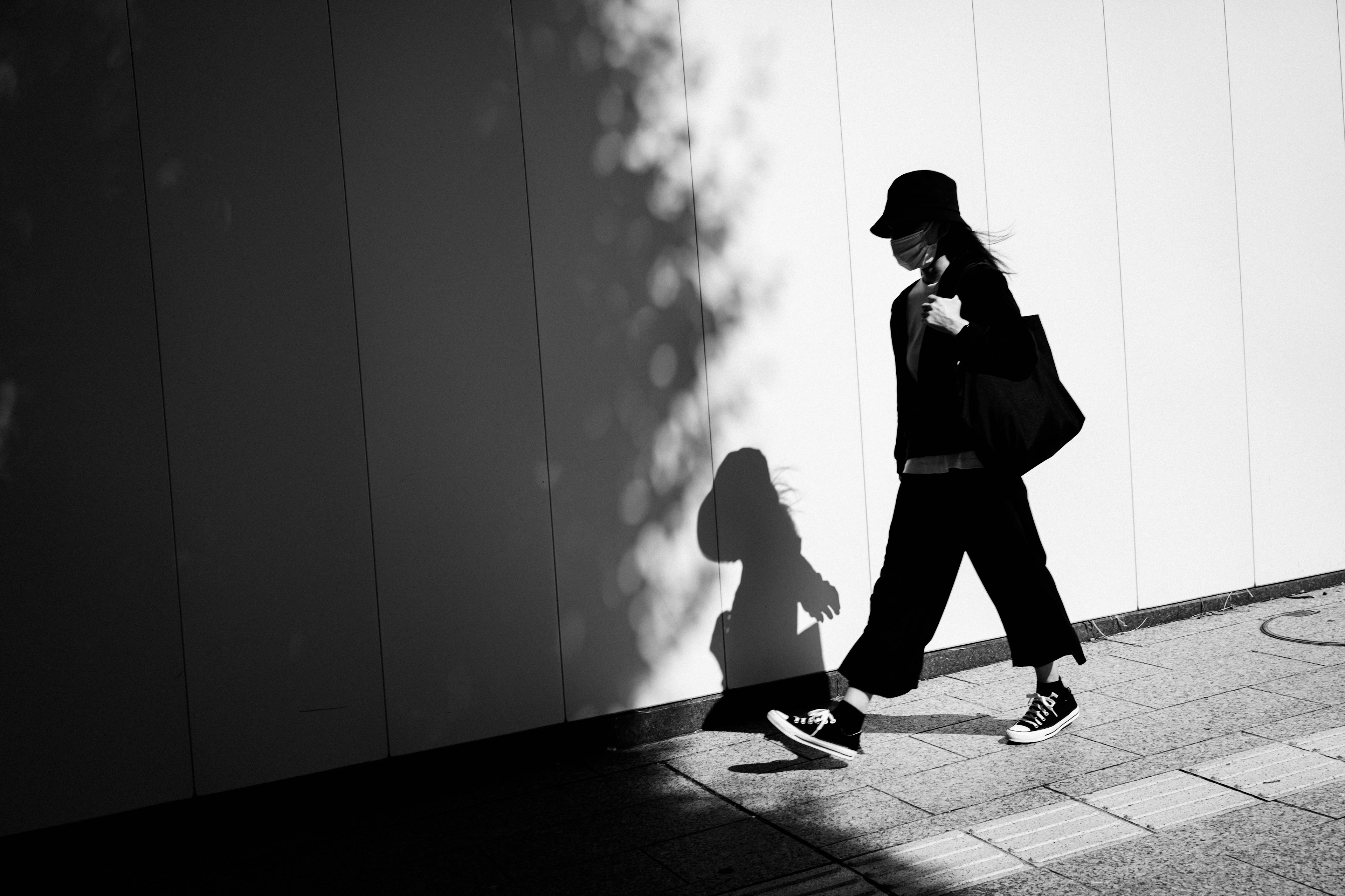 Silhouette of a woman walking in front of a white wall wearing black clothing and a hat carrying a bag